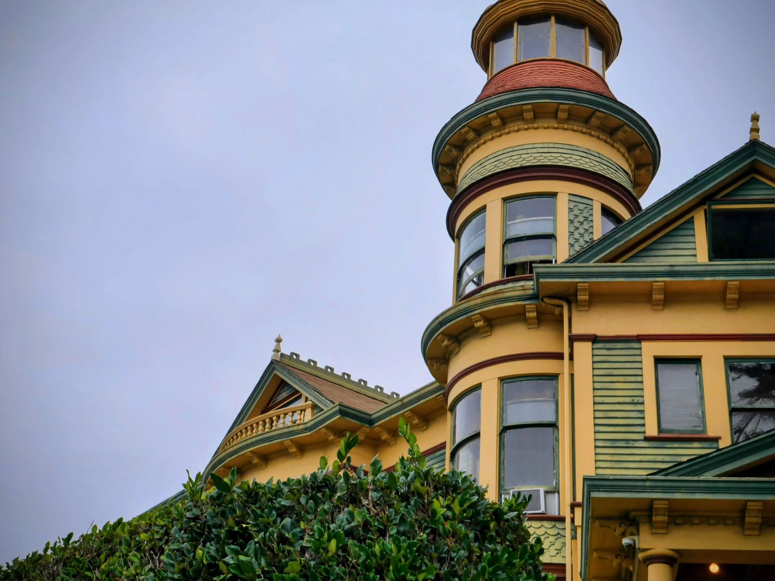 Close-up of a Victorian-style house with ornate architectural details, yellow walls, green accents, and a turret with large windows against a cloudy sky.