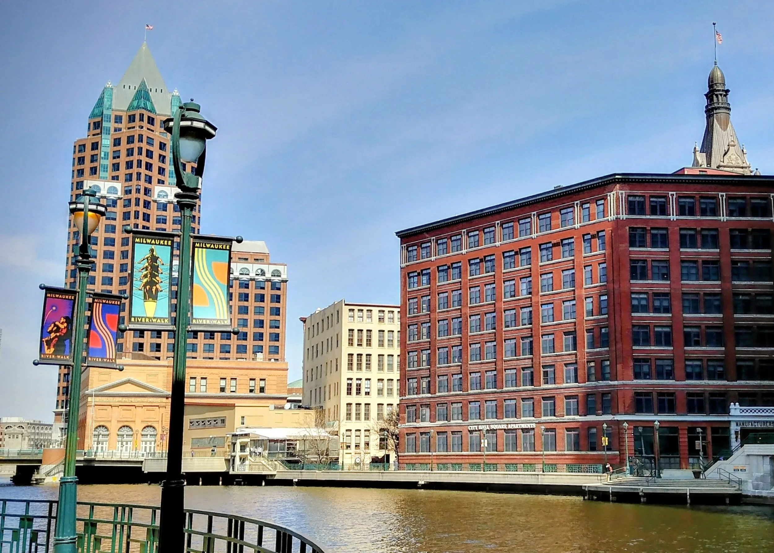 Cityscape of Milwaukee with a river in the foreground, modern and historic buildings, and decorative banners along the riverwalk.