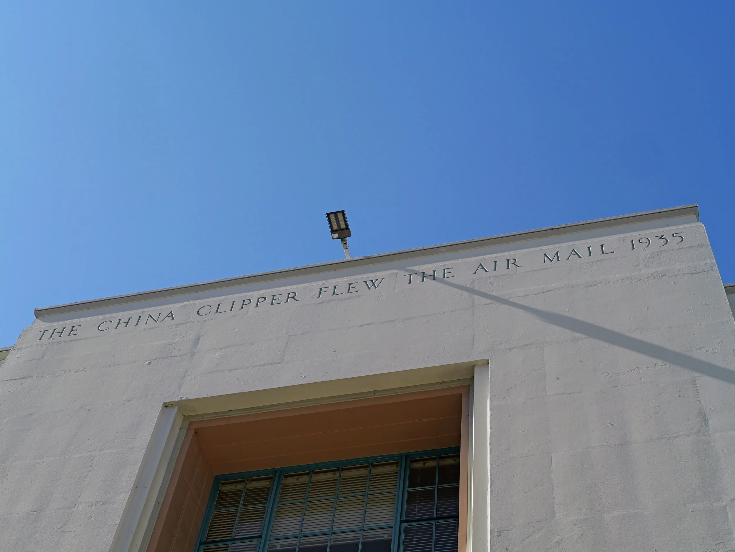 Close-up of a white building with a plaque and a large window, with a clear blue sky in the background, highlighting a tribute inscription on the building.