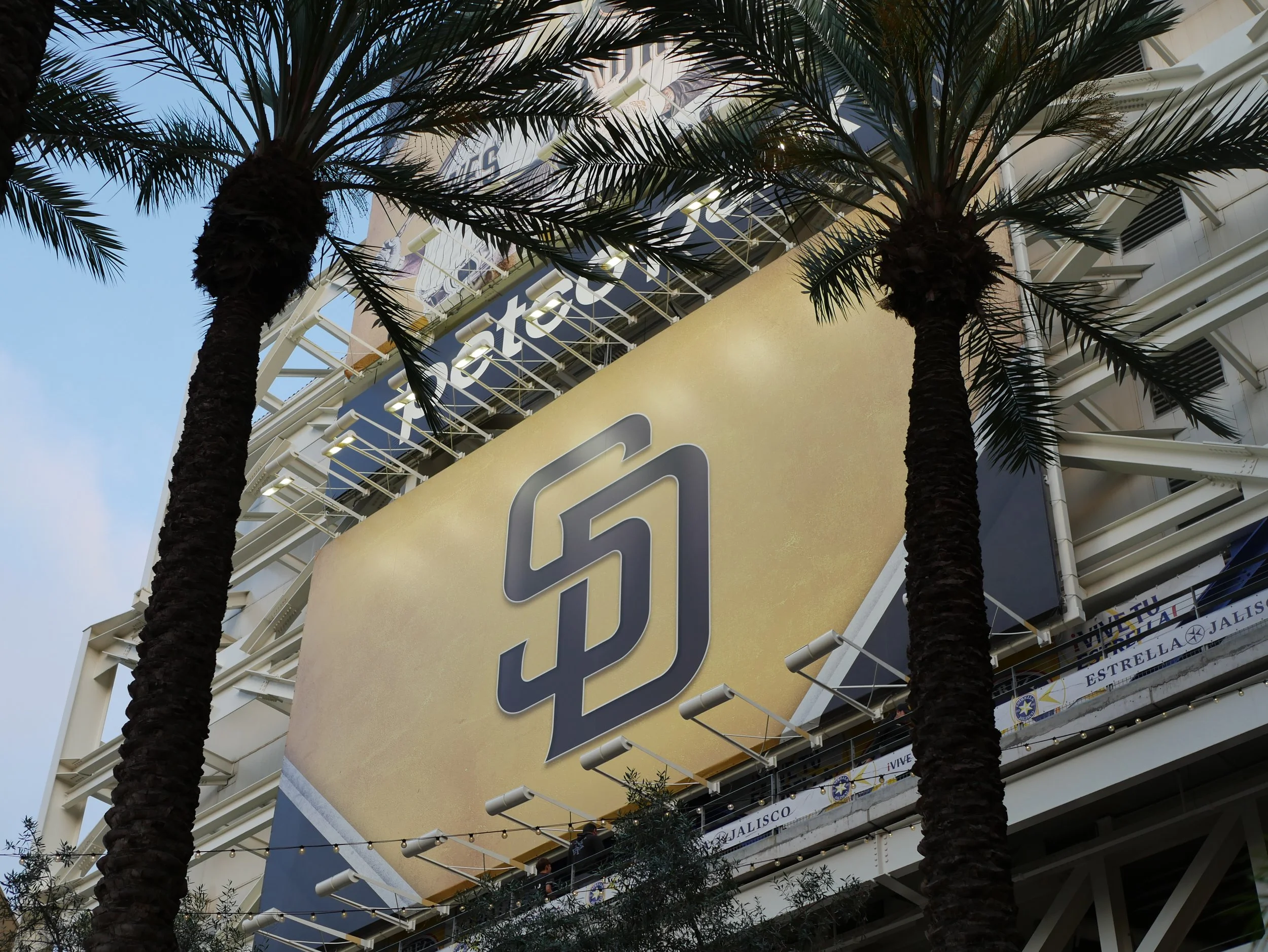 Tall palm trees in front of a large shopping mall building with a prominent gold sign and multiple floors visible, outdoors in daytime.