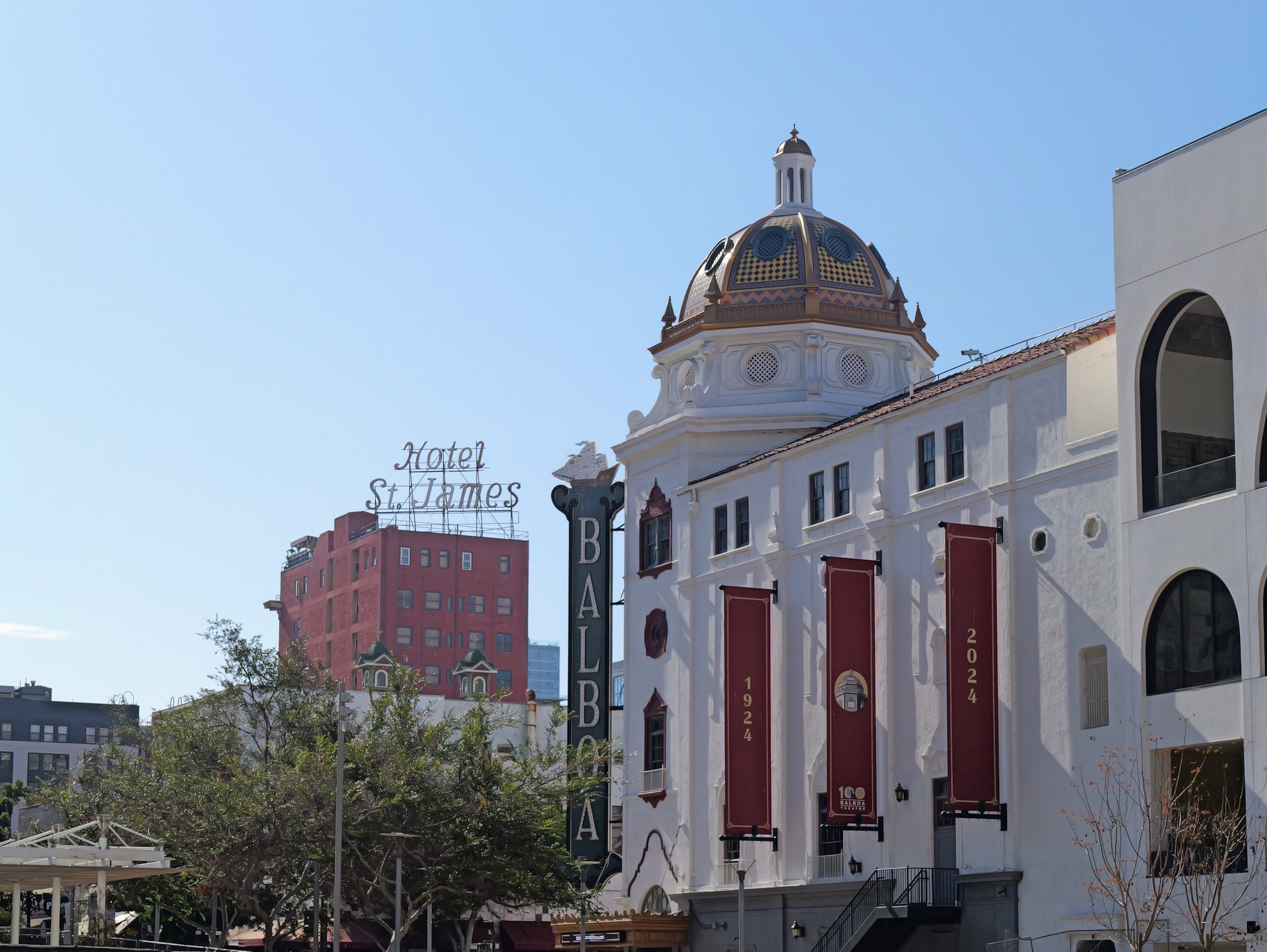 Street view of buildings with a white building featuring red banners and a decorative dome, hotel signs, and trees in the foreground with a clear blue sky in the background.