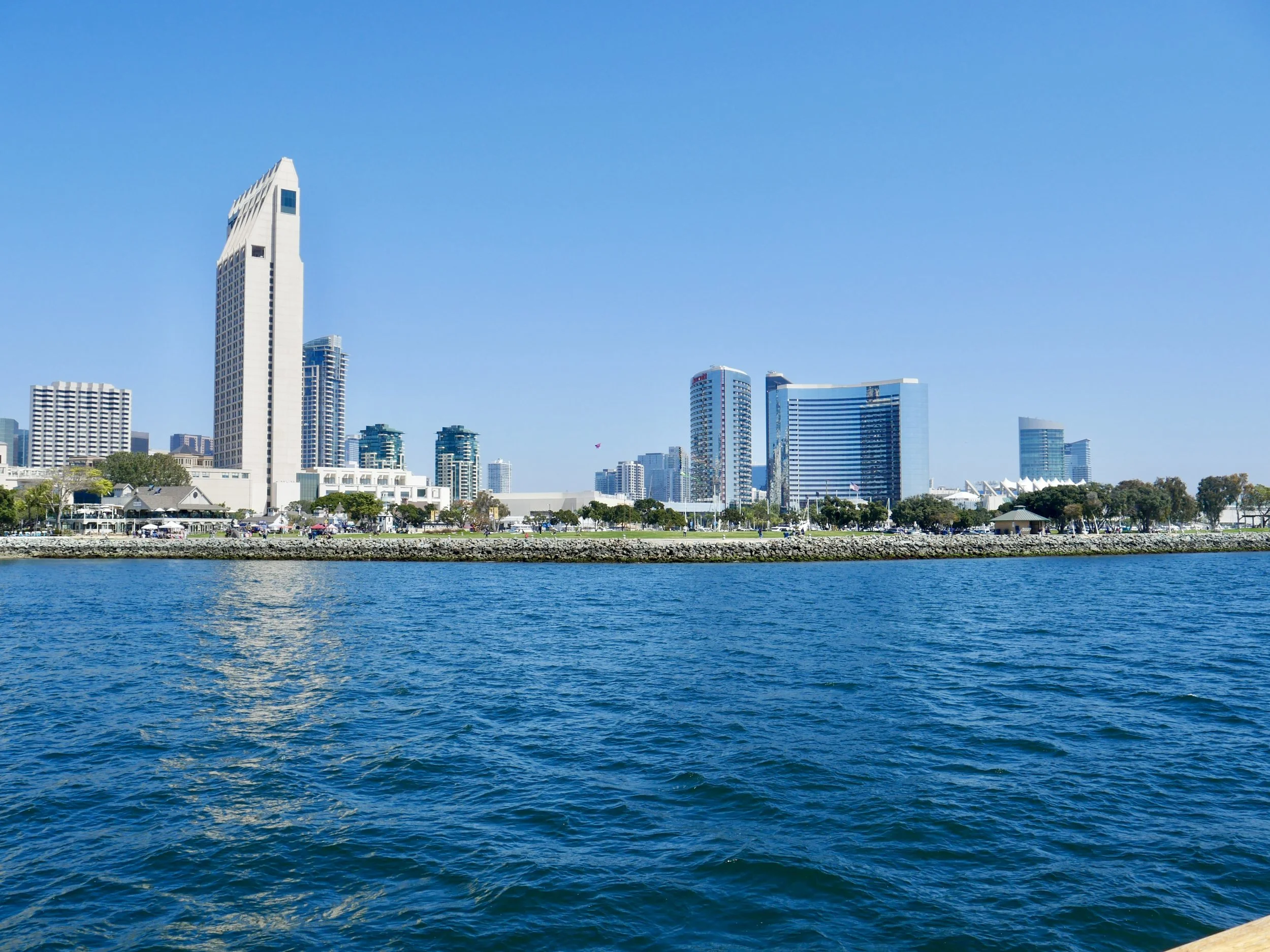 View of a city skyline with tall modern skyscrapers along the waterfront, blue sky, and water in the foreground.