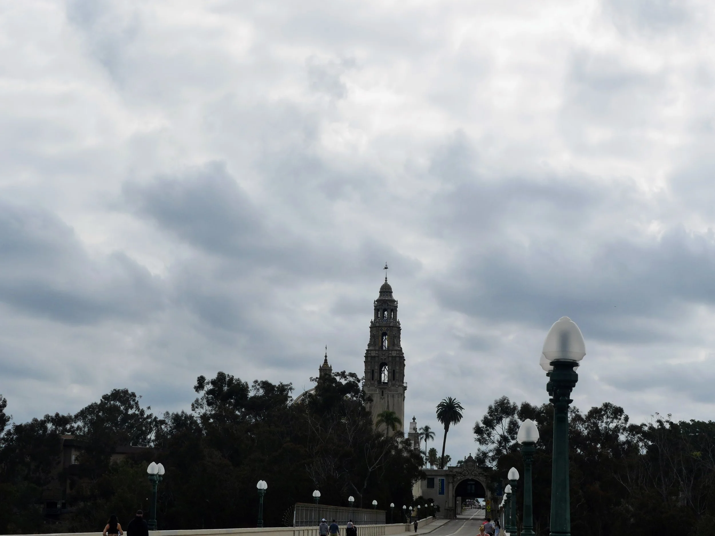 A cloudy sky over a park with a historic-style tower, trees, and outdoor lamps along a pathway with people walking.