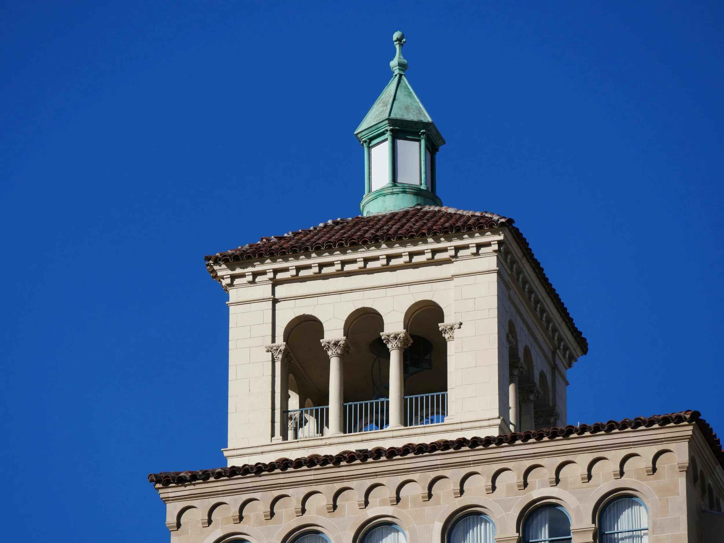 The top portion of a beige building with arched windows and decorative architectural details, topped with a roof with red tiles and a tower with a green spire against a clear blue sky.