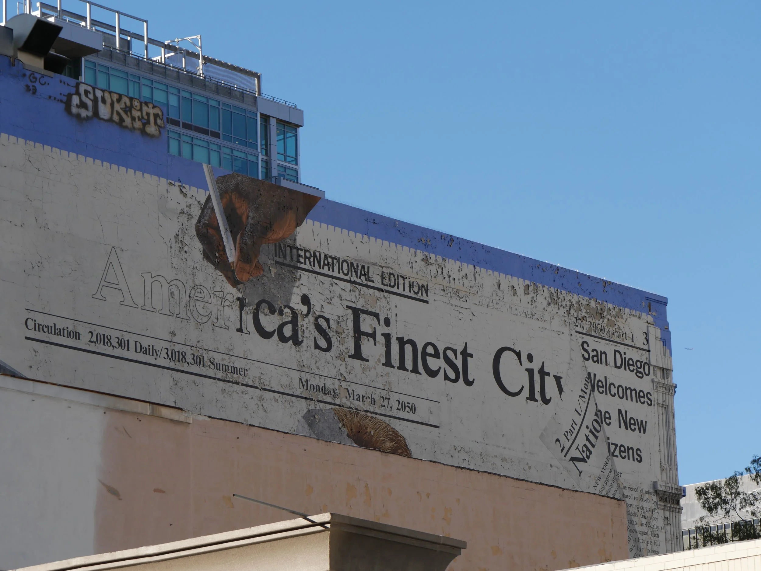 A cityscape showing a large billboard with text about America's Finest City, San Diego, with a modern glass building behind it, under a clear blue sky.