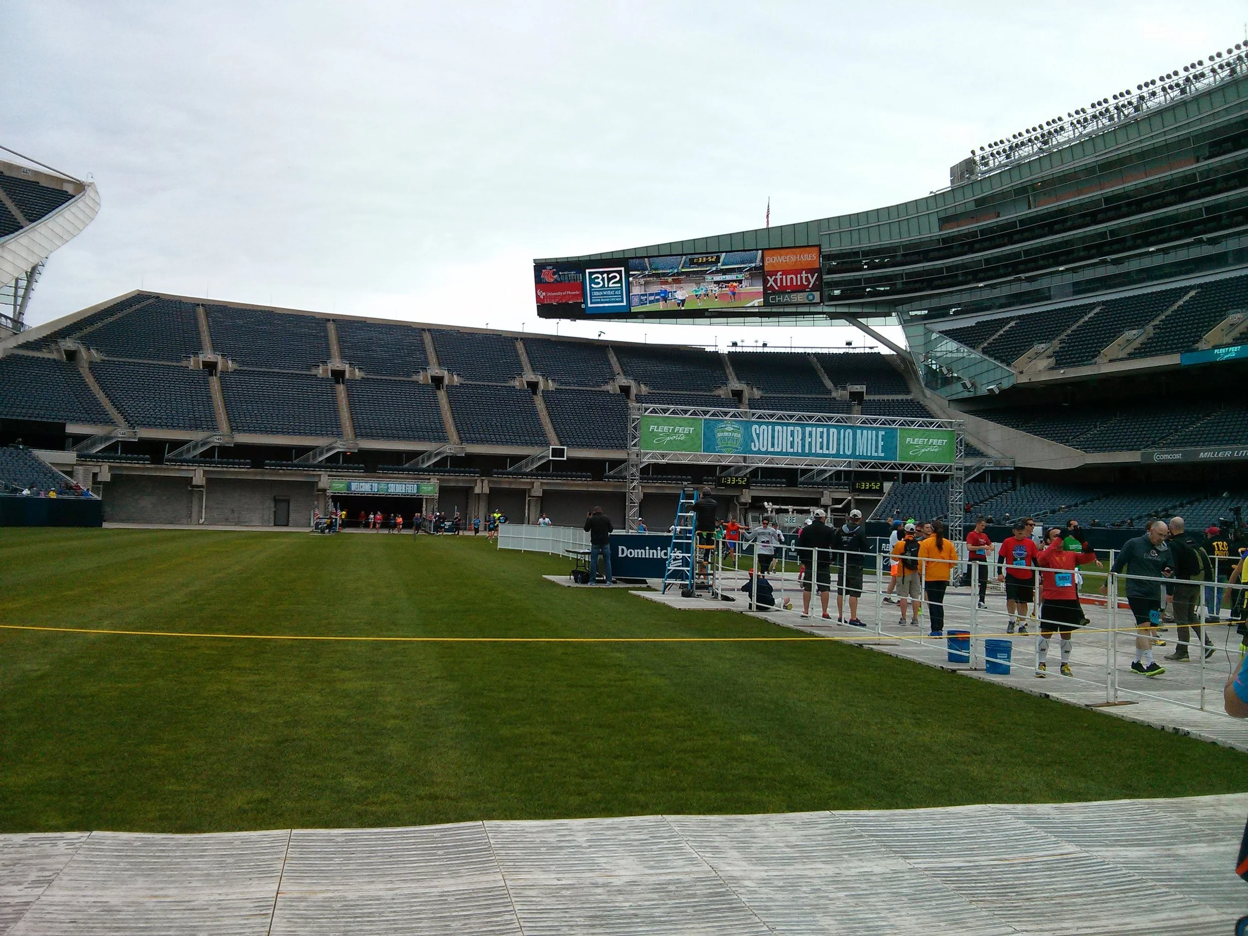 View of Soldier Field stadium with runnings event setup, including a large digital scoreboard, tents, and a group of runners and volunteers on the field.