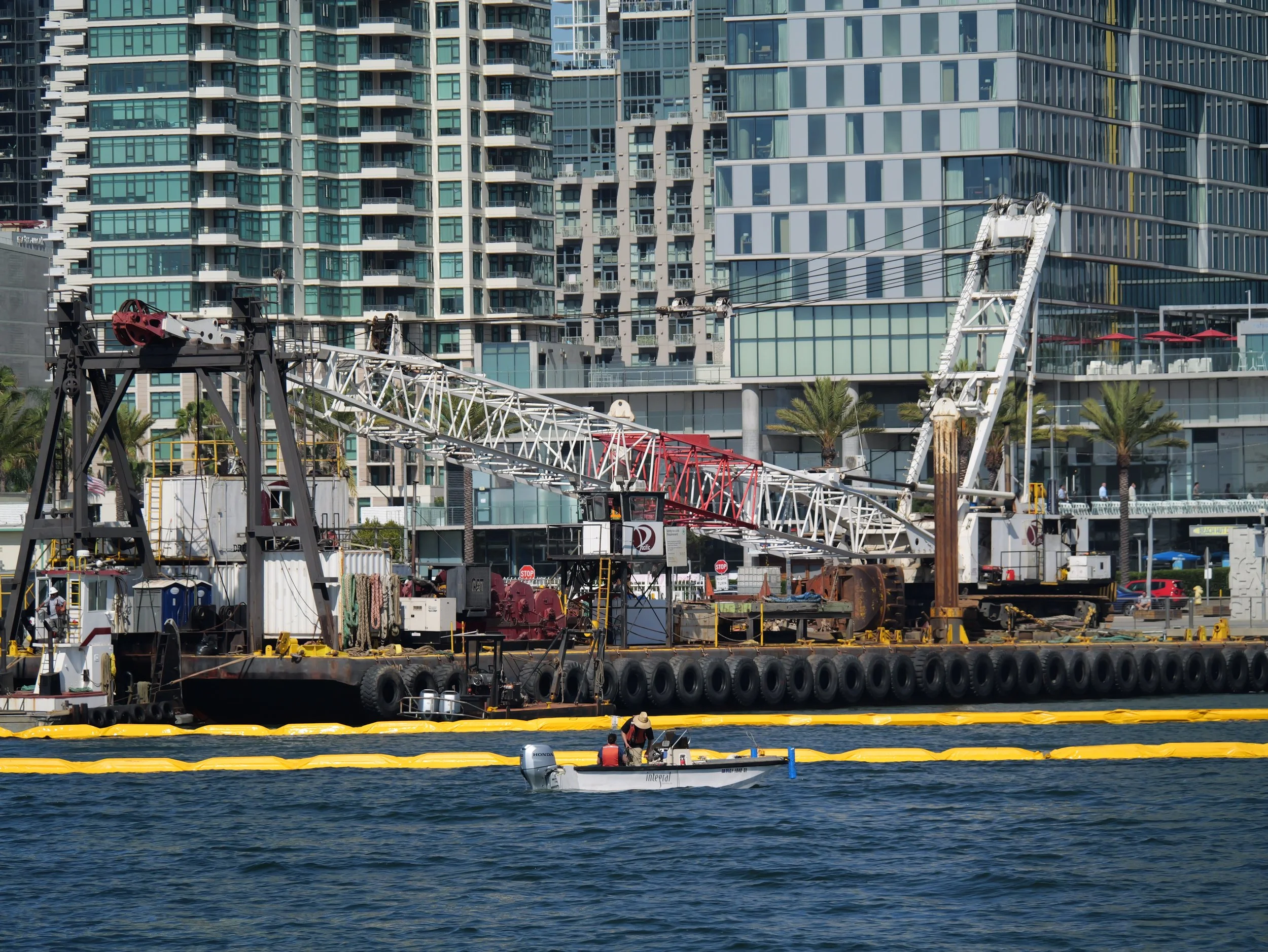 A city waterfront scene with modern high-rise buildings in the background, a boat on the water carrying two people, and part of a construction site or dock with equipment and yellow safety barriers in the foreground.