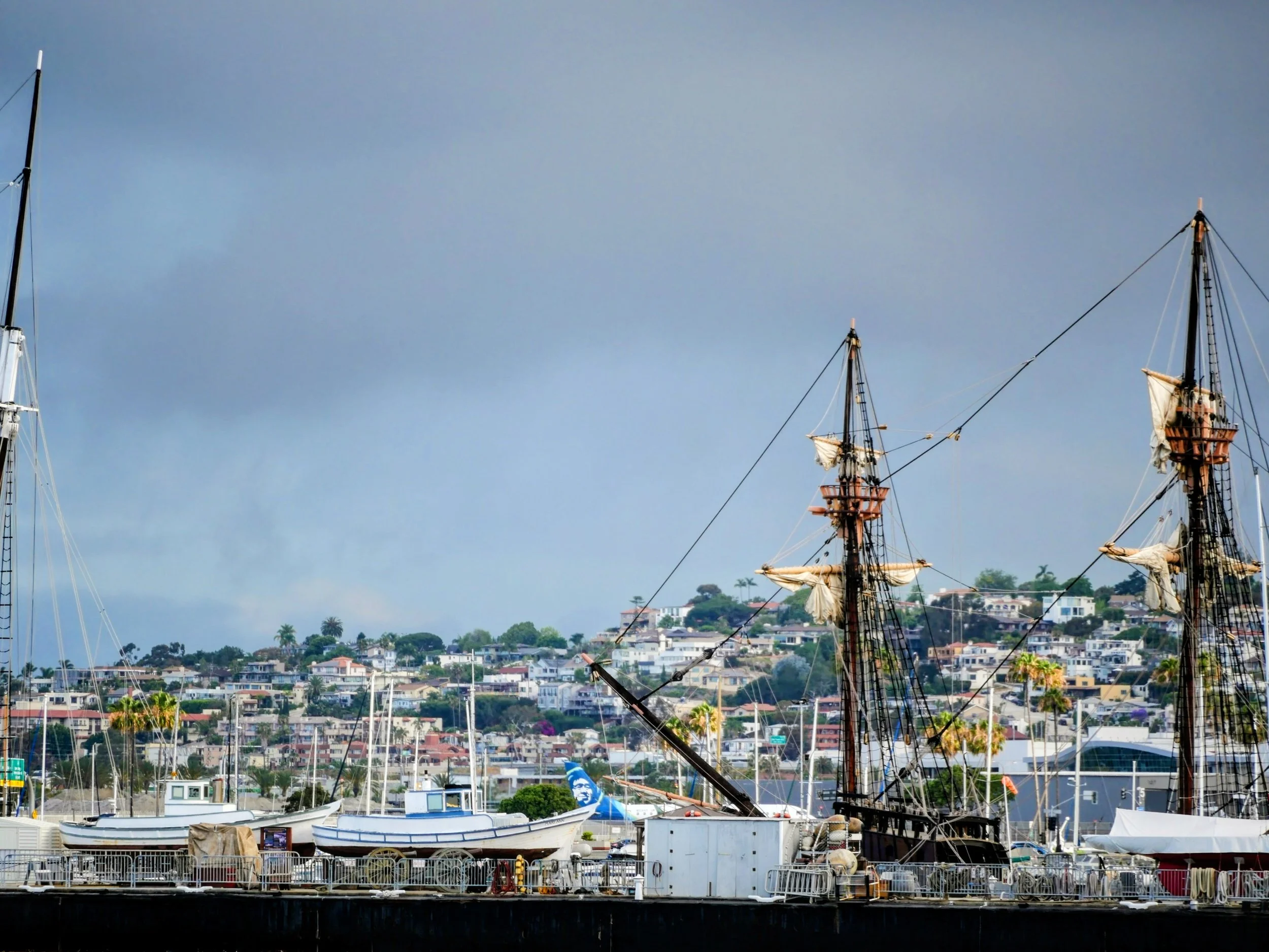 A harbor scene with two old-style sailing ships with tall masts and a row of modern boats docked along the pier. In the background, a hillside filled with houses and greenery under a cloudy sky.