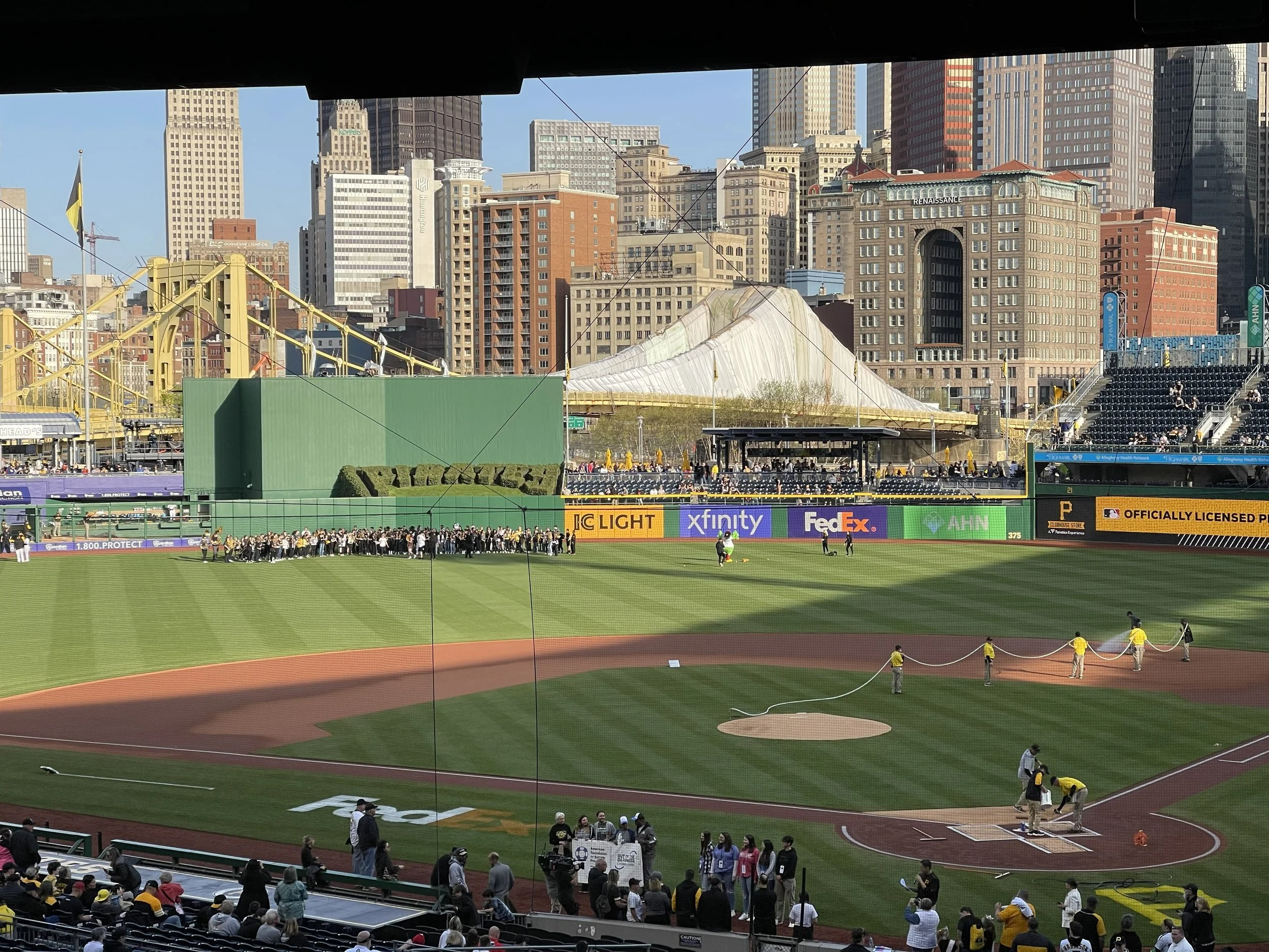 Baseball field with grounds crew preparing the infield, spectators in stands, and city skyline in the background.