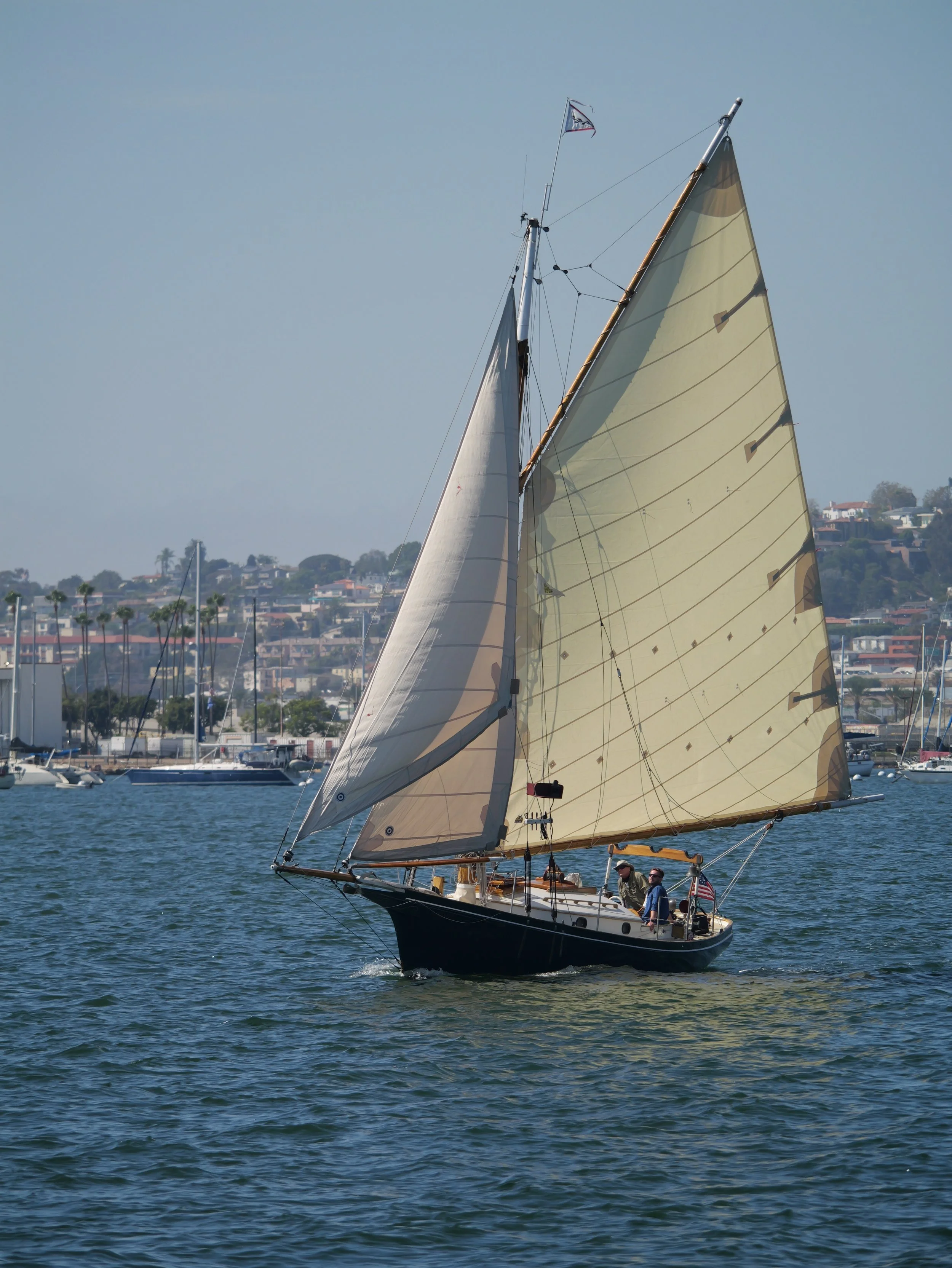 A sailboat with two people on board, sailing on a body of water with a cityscape and hillside in the background. The boat has large beige sails and a small flag at the top of the mast.