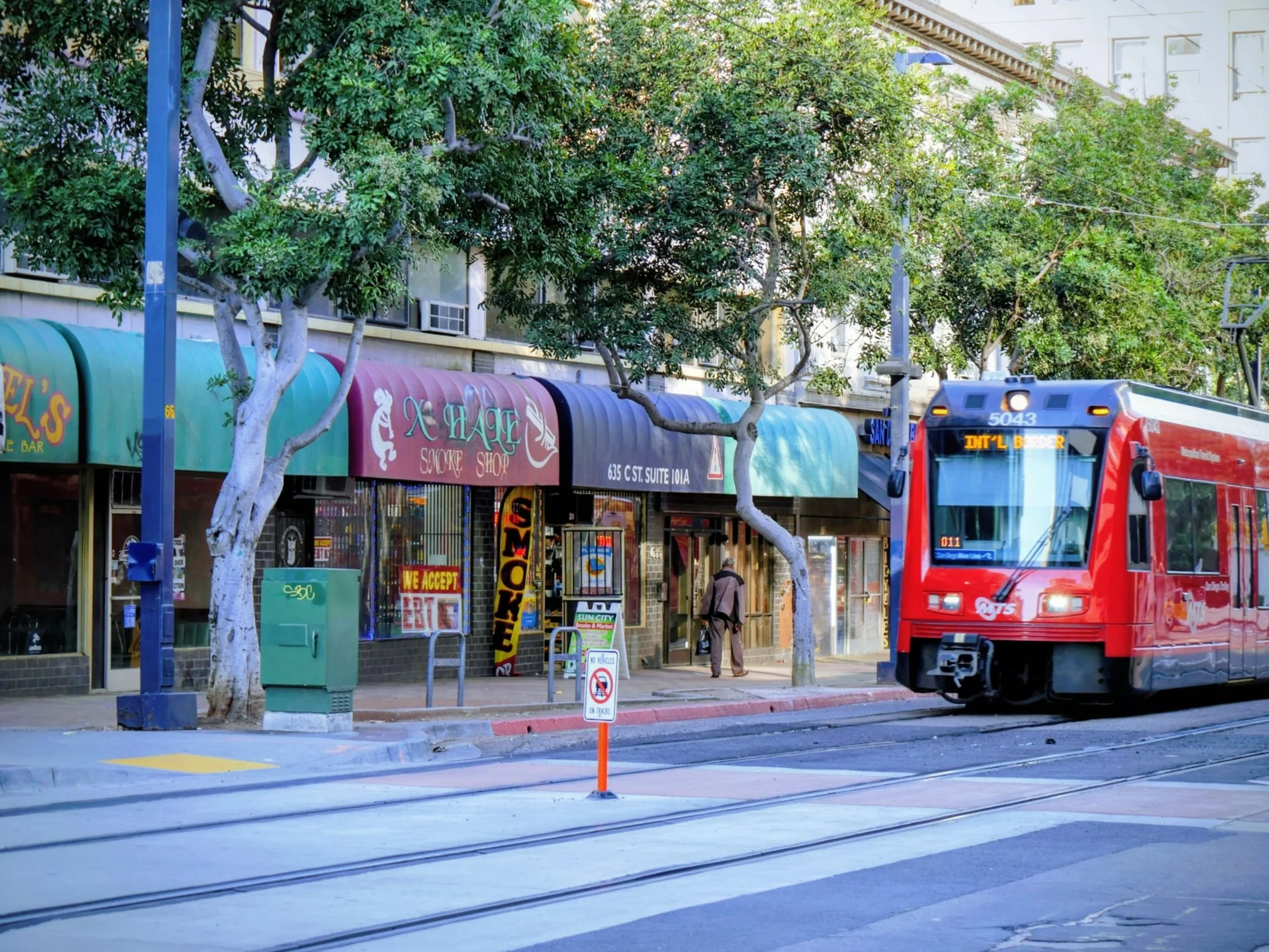 Street scene with shops, trees, a red streetcar, and pedestrians.