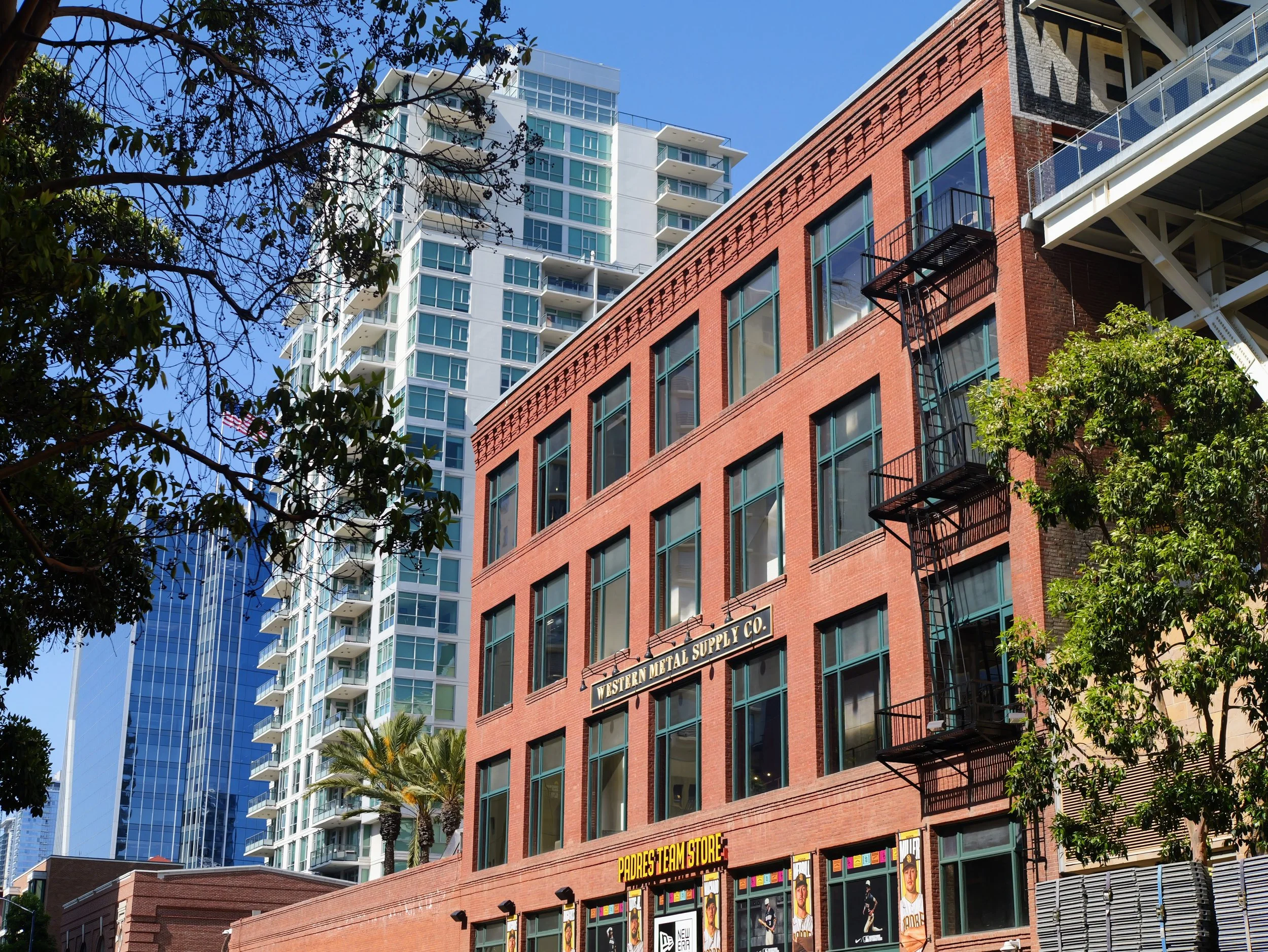 Tall modern white high-rise building with glass balconies in the background, and a red brick building with large windows and exterior fire escape in the foreground, with trees and a clear blue sky.