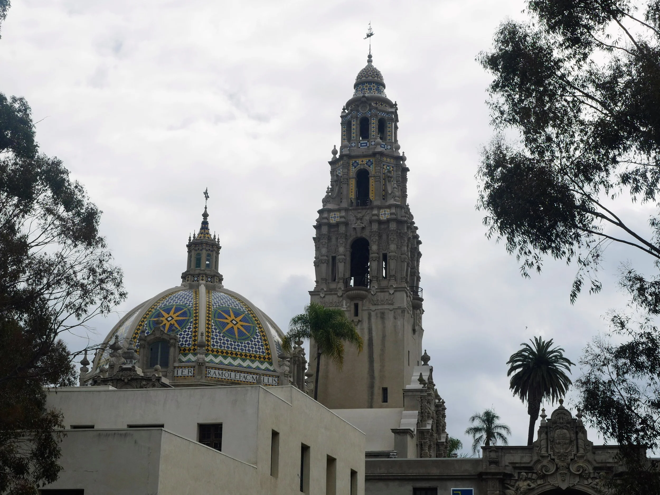 View of a historical building with a clock tower and a colorful dome, partially obscured by trees, under an overcast sky.