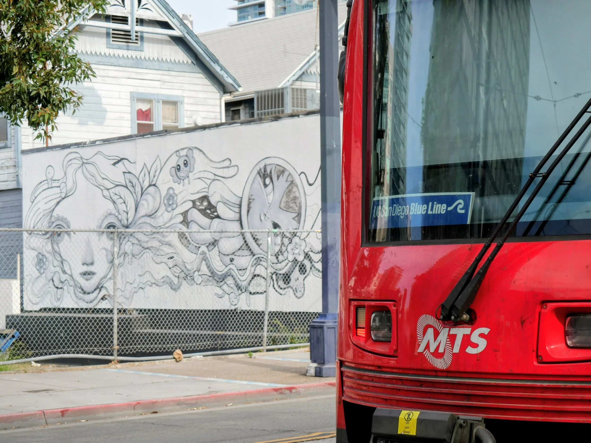 A red vehicle with the logo MTS and text 'Los Angeles Blue Line' is parked on the street. In the background, there is a graffitied white wall, houses with blue trim, a tree, and a chain-link fence.