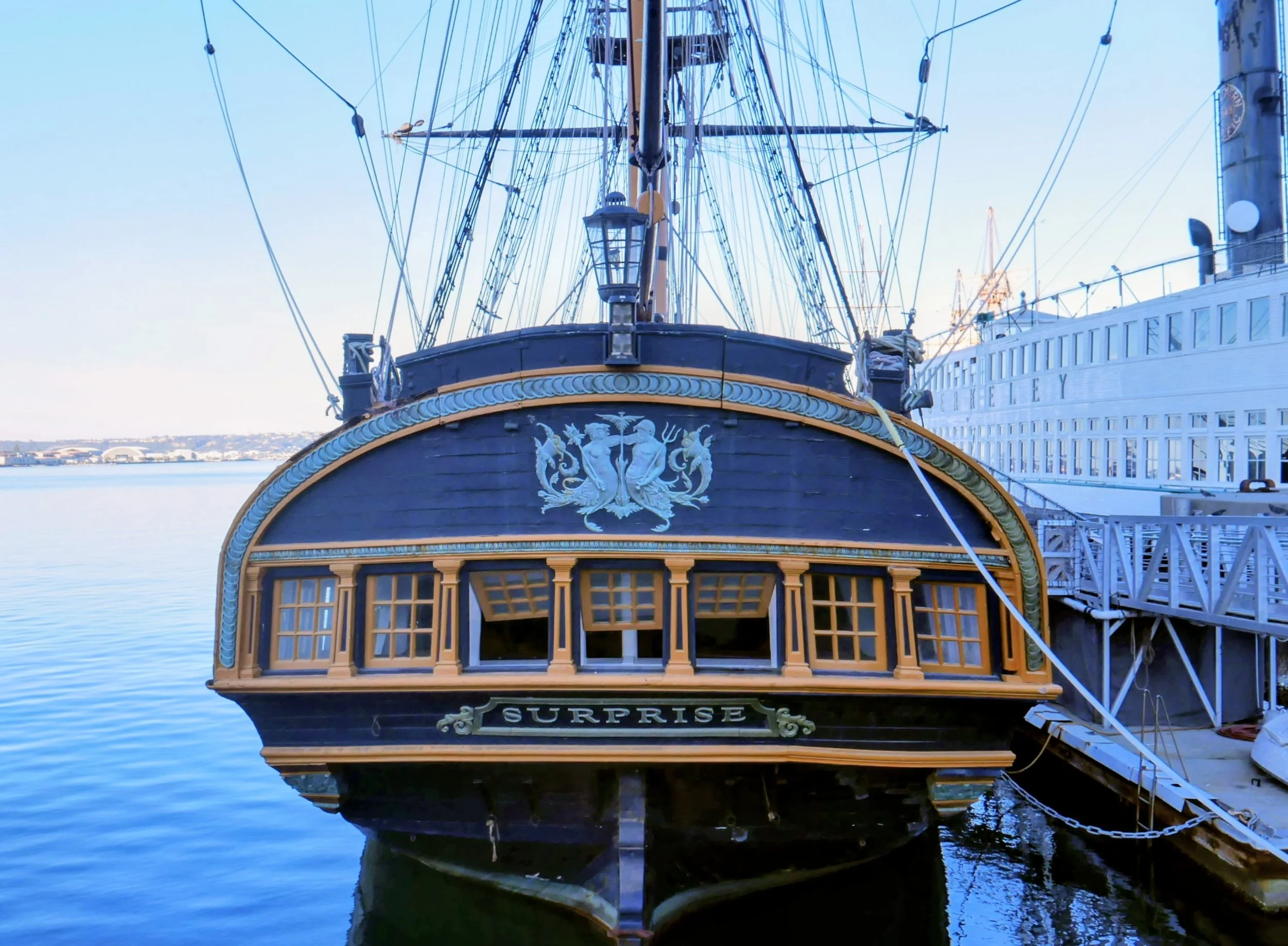 Front view of a historic sailing ship named 'Surprise' docked at a pier, with a large white cruise ship next to it, calm water, and a distant shoreline.