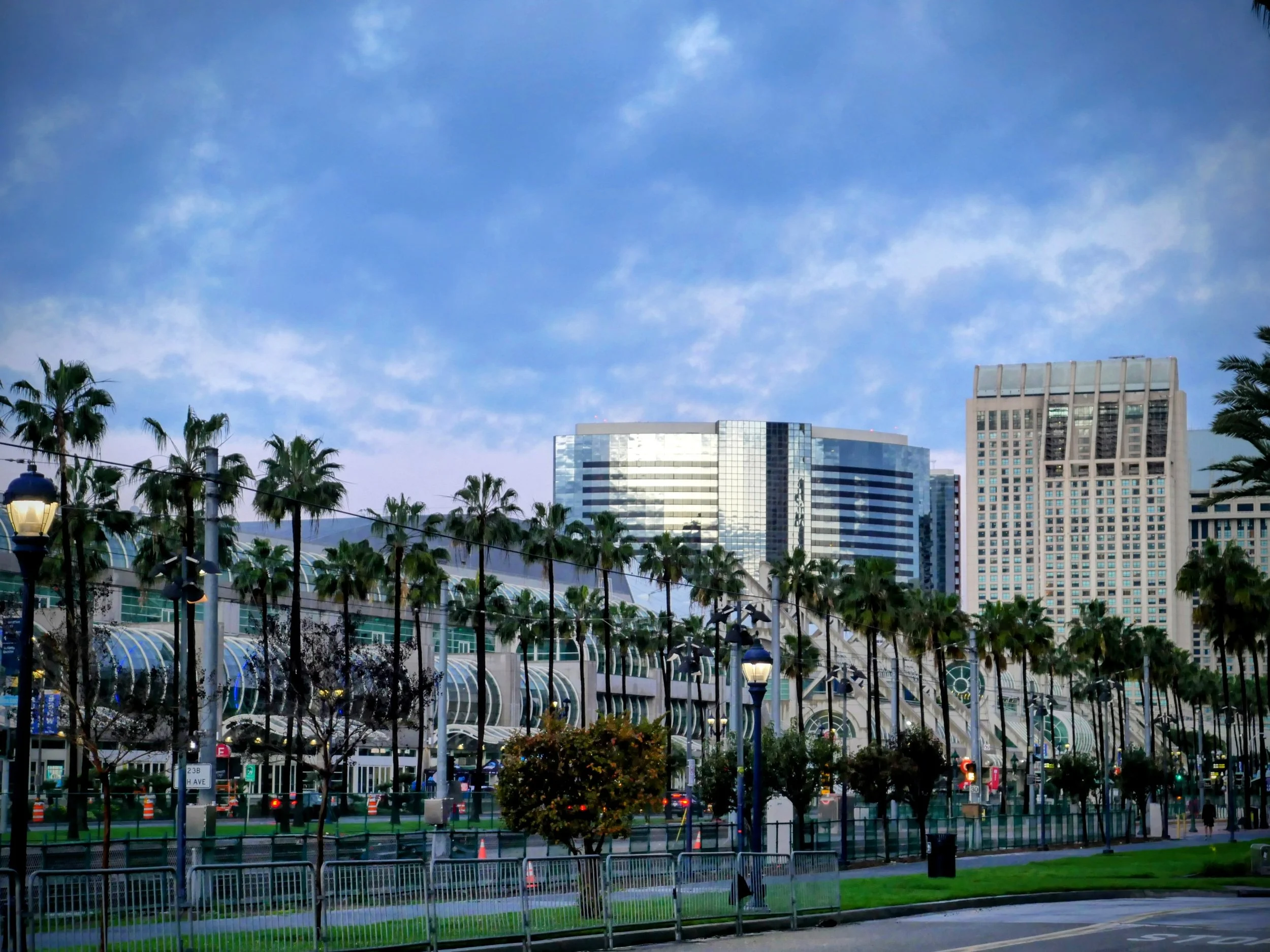 Cityscape with modern glass buildings, palm trees, street lamps, traffic cones, and people walking in the foreground under a partly cloudy sky.