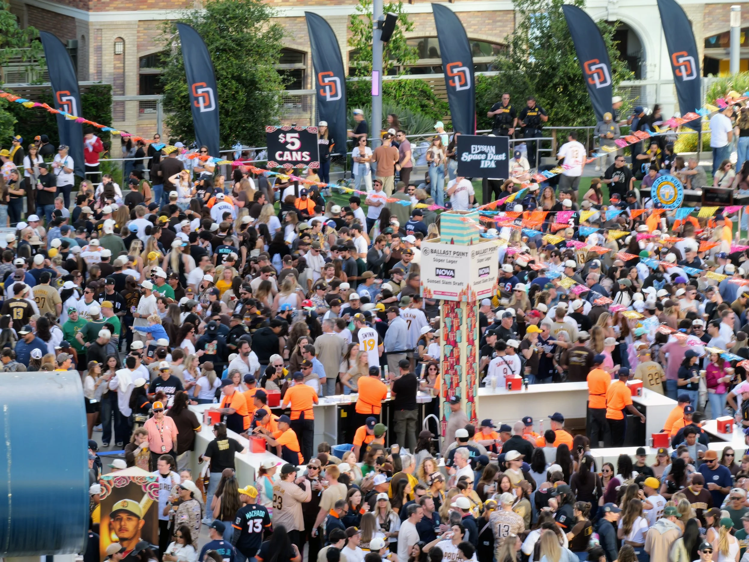 A crowded outdoor event with many people, some wearing sports jerseys and hats. There are black flags with orange and white logos, colorful decorations hanging across, and several signs advertising food and beverages. The background shows a building 