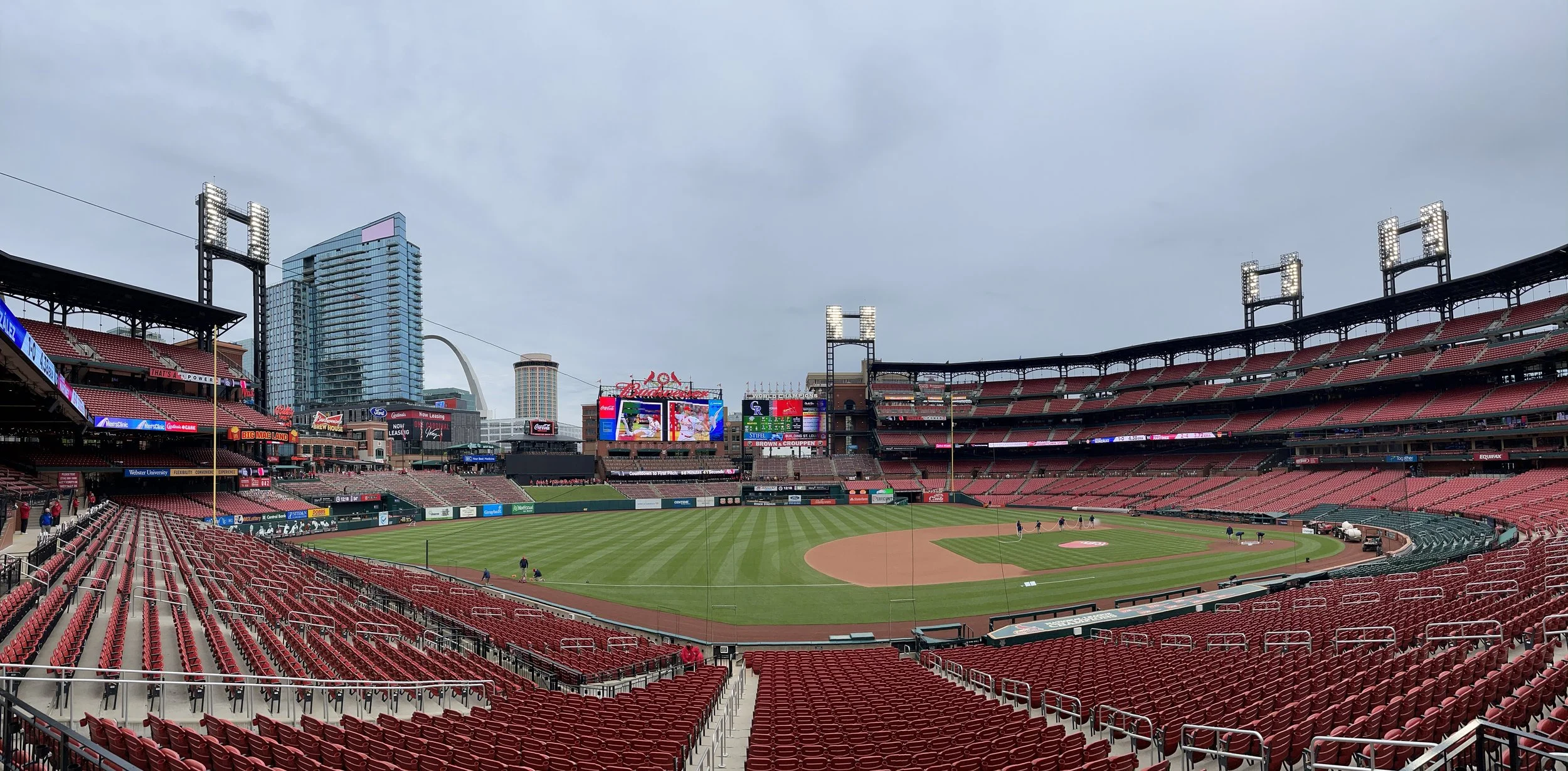 Empty baseball stadium with red seats, green field, and city skyline in the background on a cloudy day.