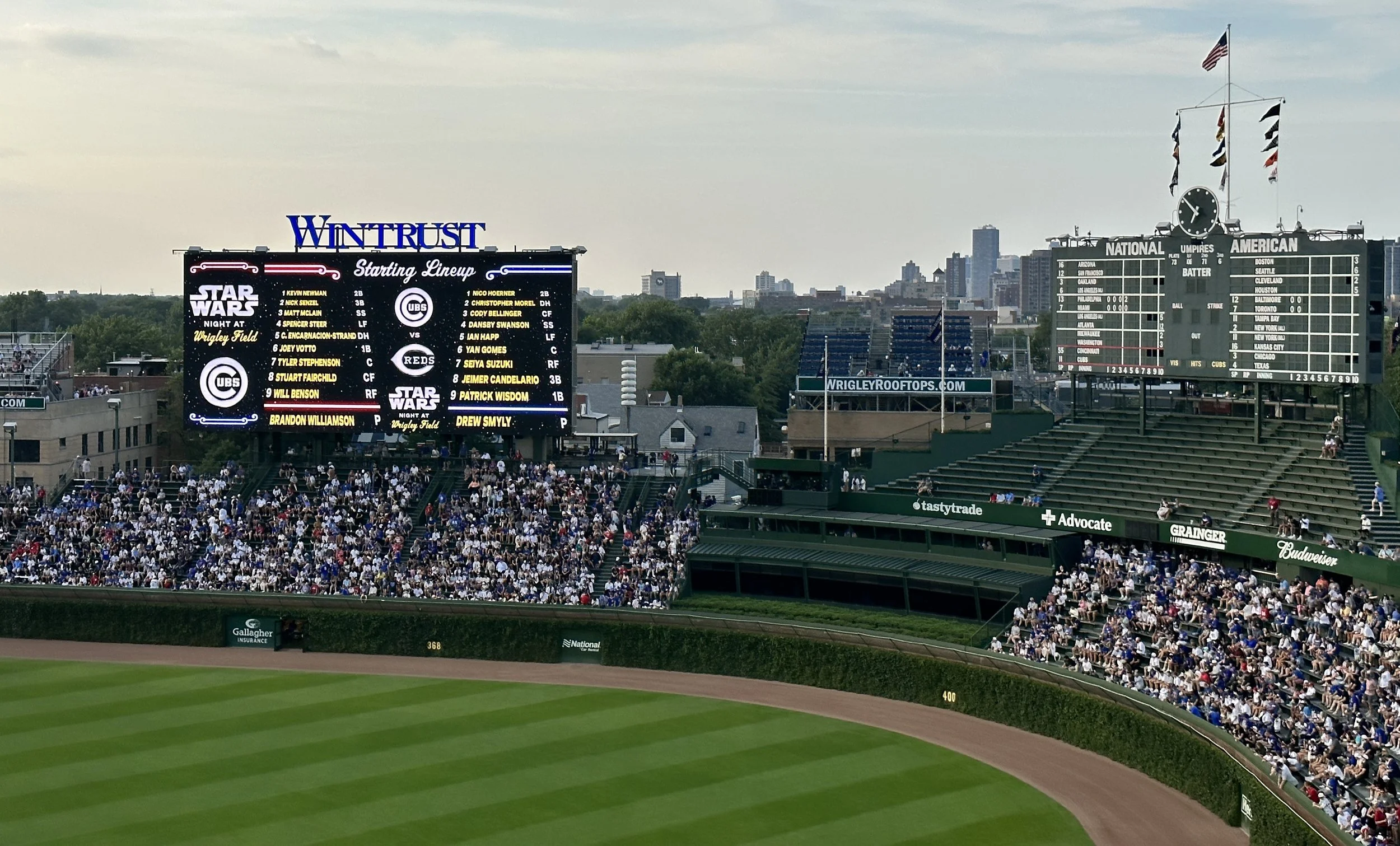 A baseball stadium filled with spectators, showing a large scoreboard and seating area, with the city skyline in the background.