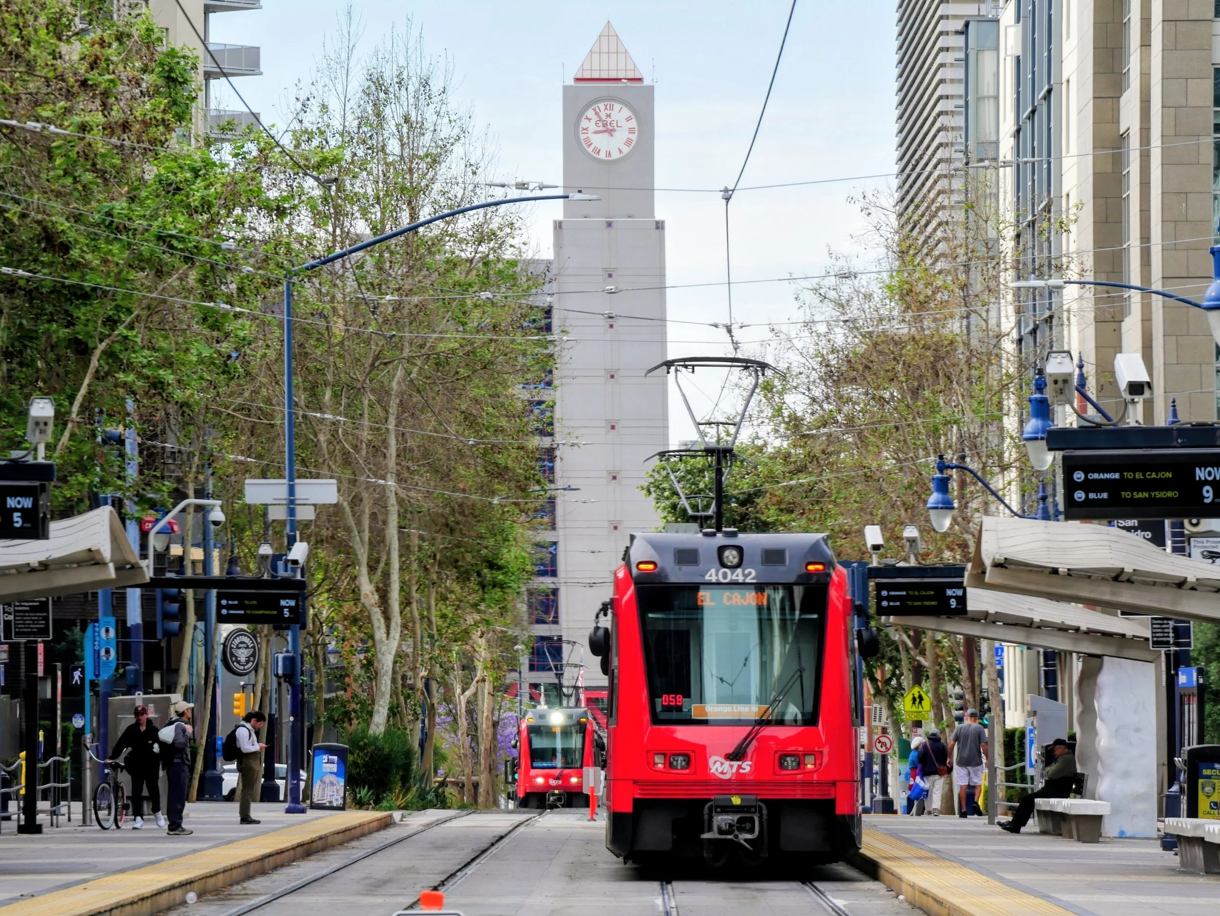 View of a city street with a red trolley bus on tracks, people walking and waiting, traffic signals, and a tall clock tower in the background.