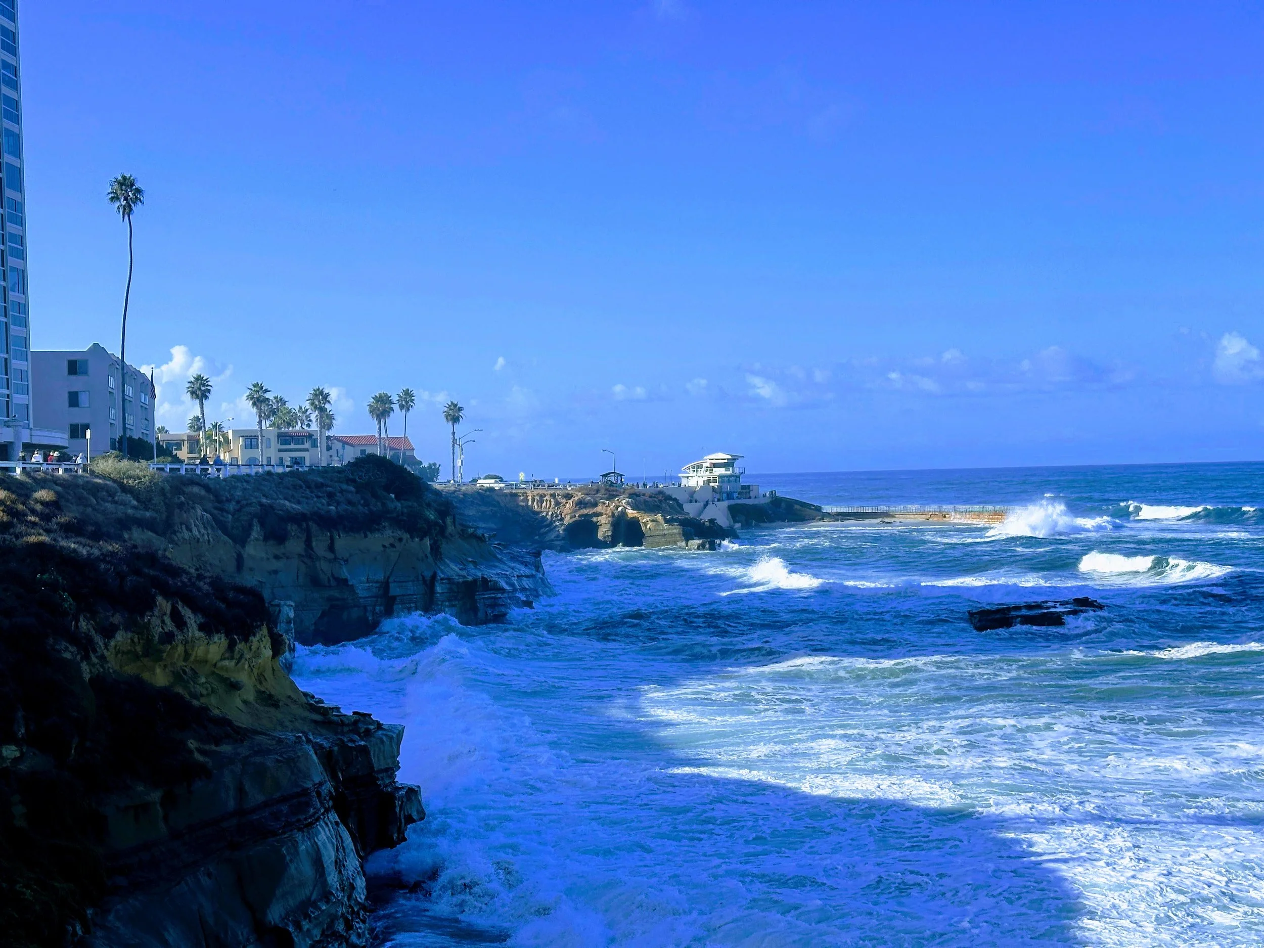 A coastal scene with high cliffs, a blue ocean with waves, and buildings with palm trees along the shoreline under a clear blue sky.