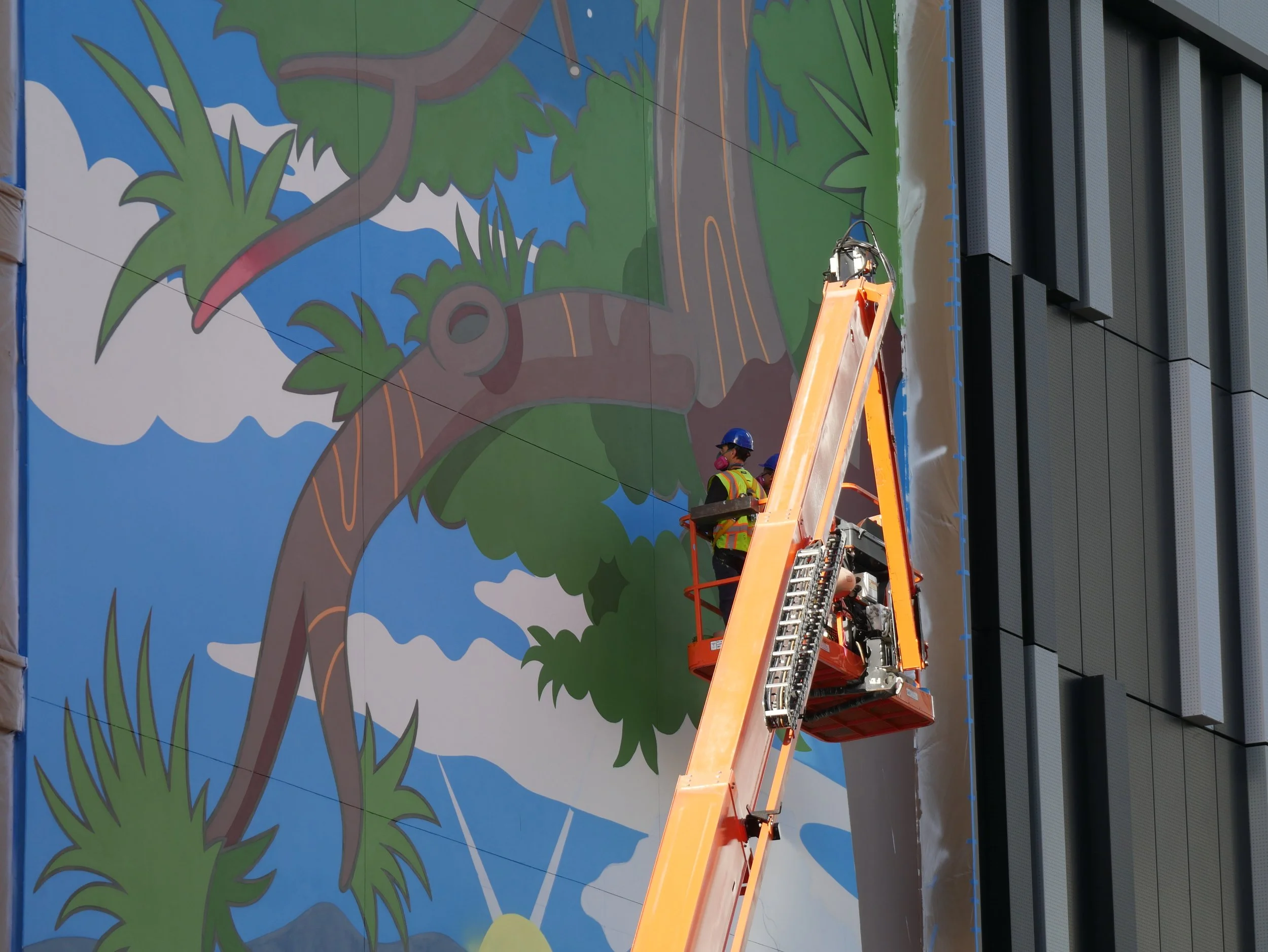 Workers on an elevated platform painting a mural of a tree with green leaves and blue sky on a building wall.