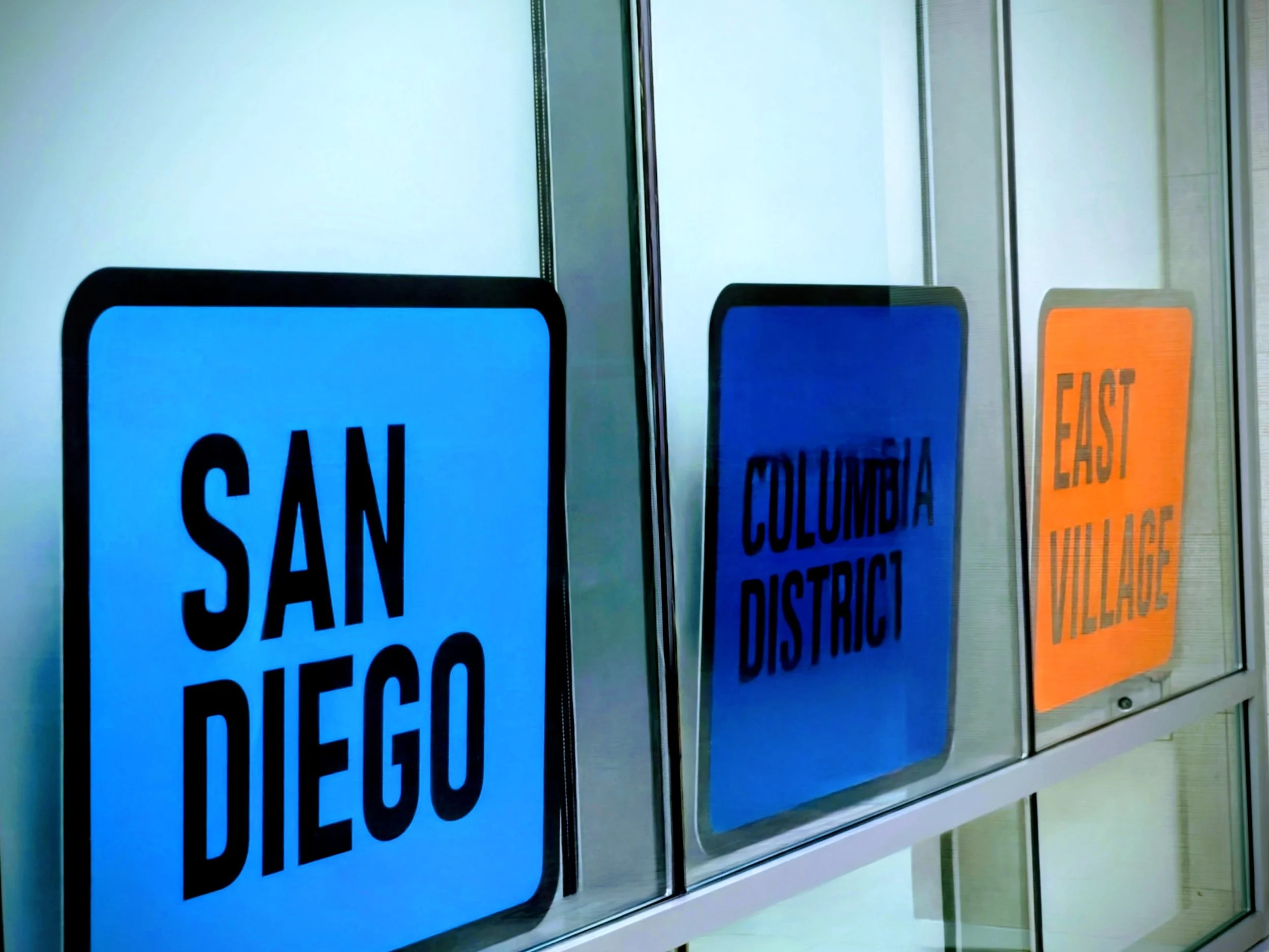 Framed signs on a wall with text including 'CANCEL' and 'EAST' in bold. The signs are arranged in a row, with reflective glass panels behind them.