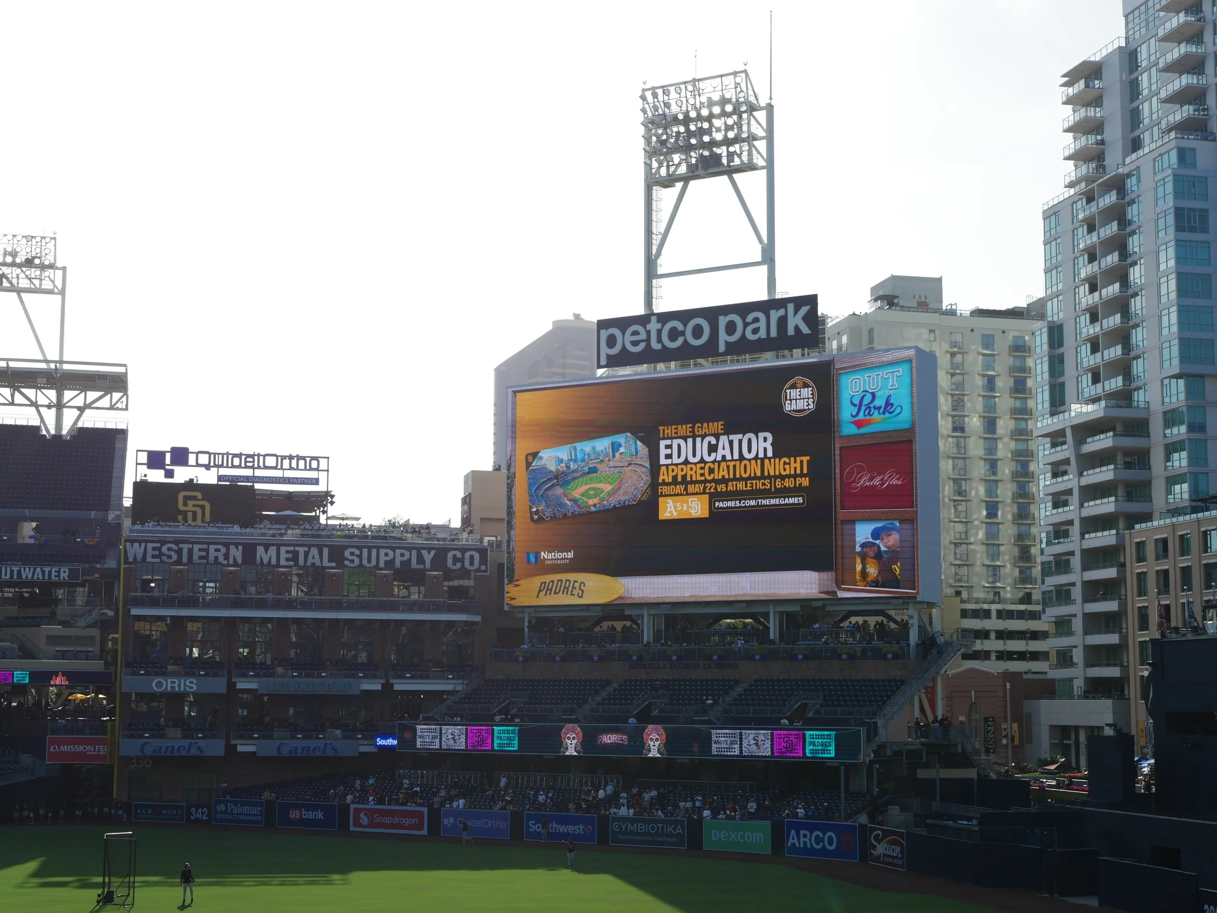 View of Petco Park stadium with advertisements, scoreboard, and surrounding residential buildings, during daytime with a clear sky.