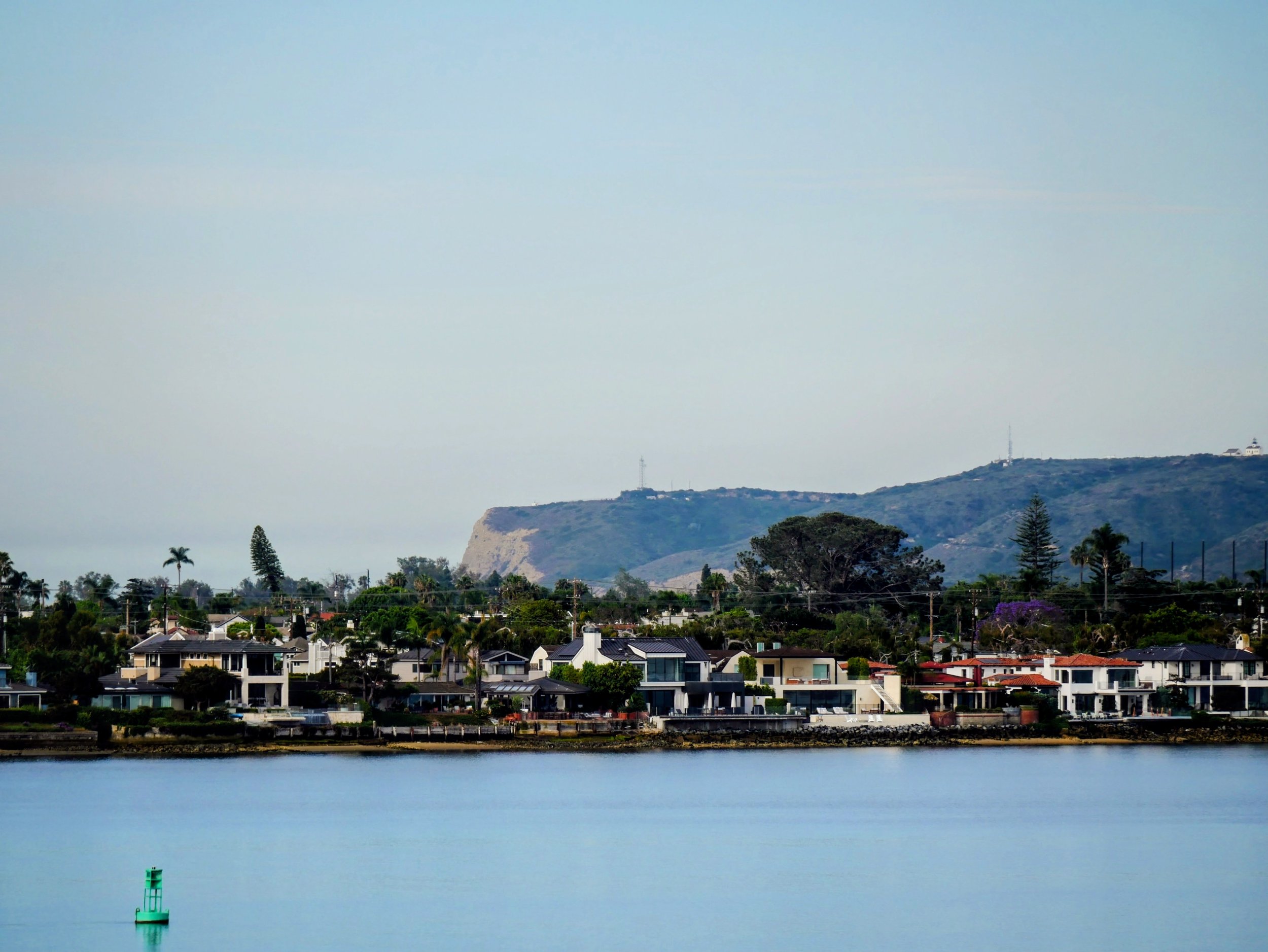 A coastal scene with calm water in the foreground, upscale houses lining the shore in the middle, and rolling hills with some structures on top in the background.