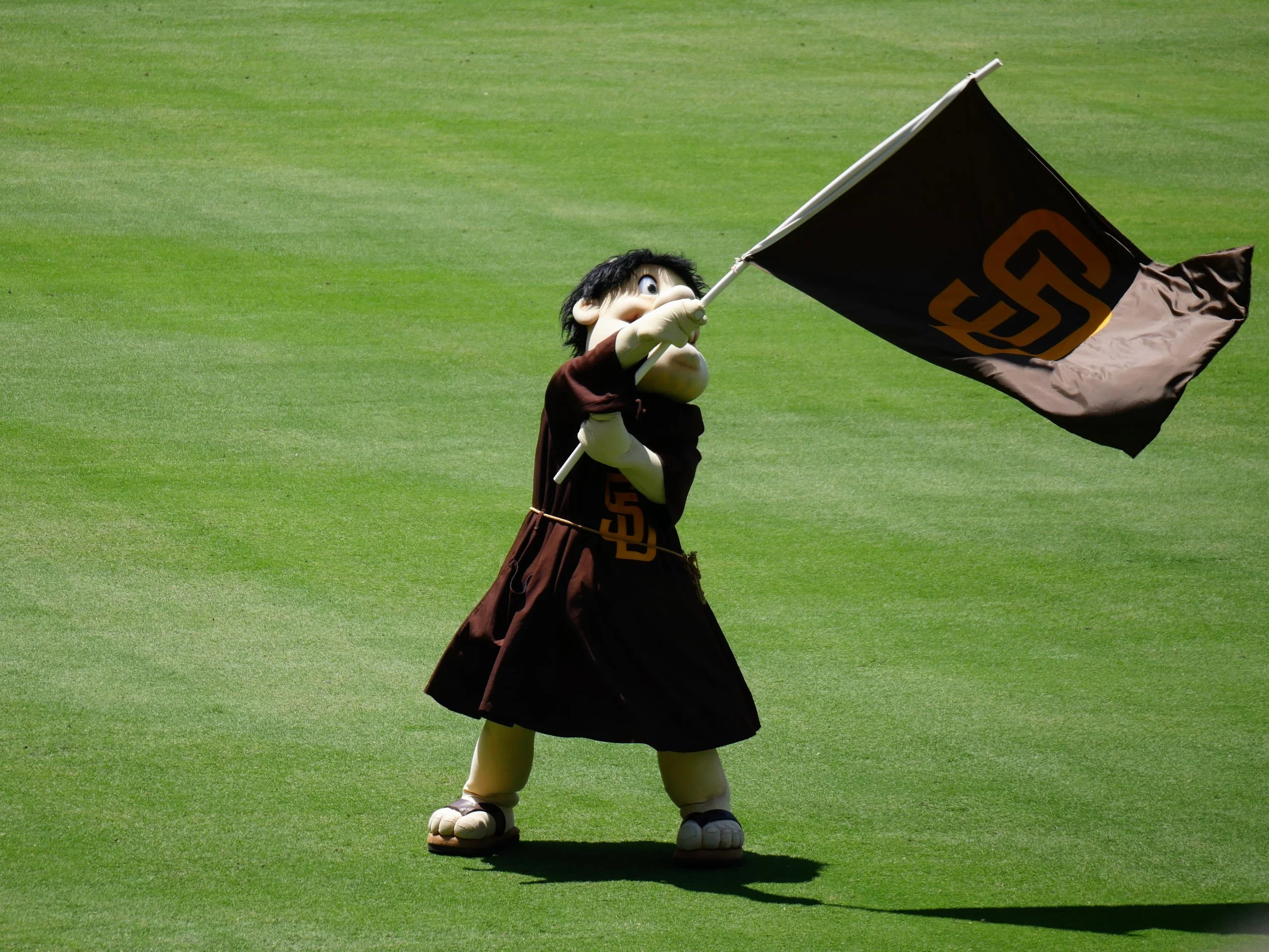 A mascot character dressed in a brown costume with a large head, black hair, and a white face, holding a black and yellow flag on a golf course with green grass.