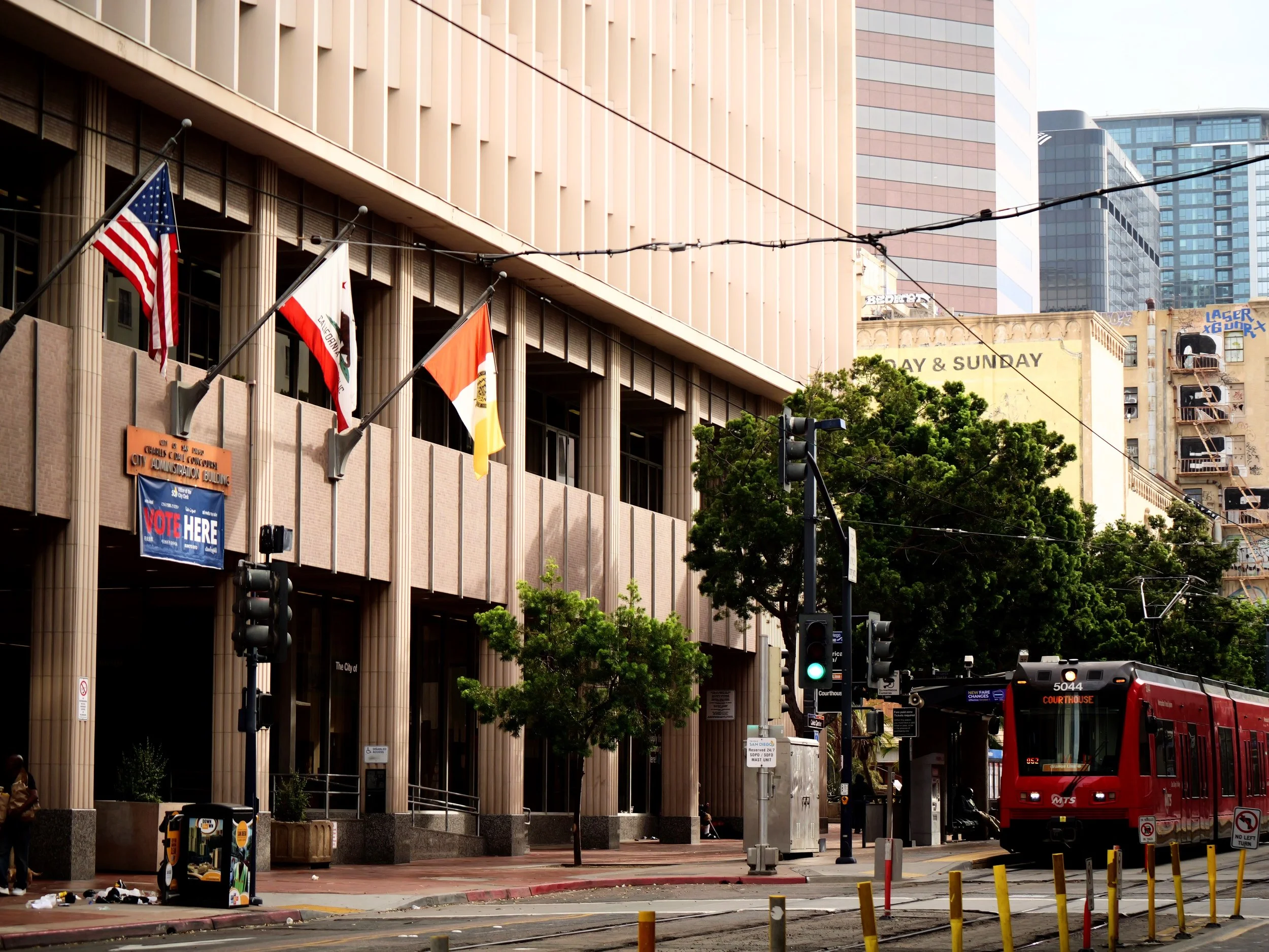 Urban street scene with a city hall or government building, flags, a red bus, traffic lights, and trees.