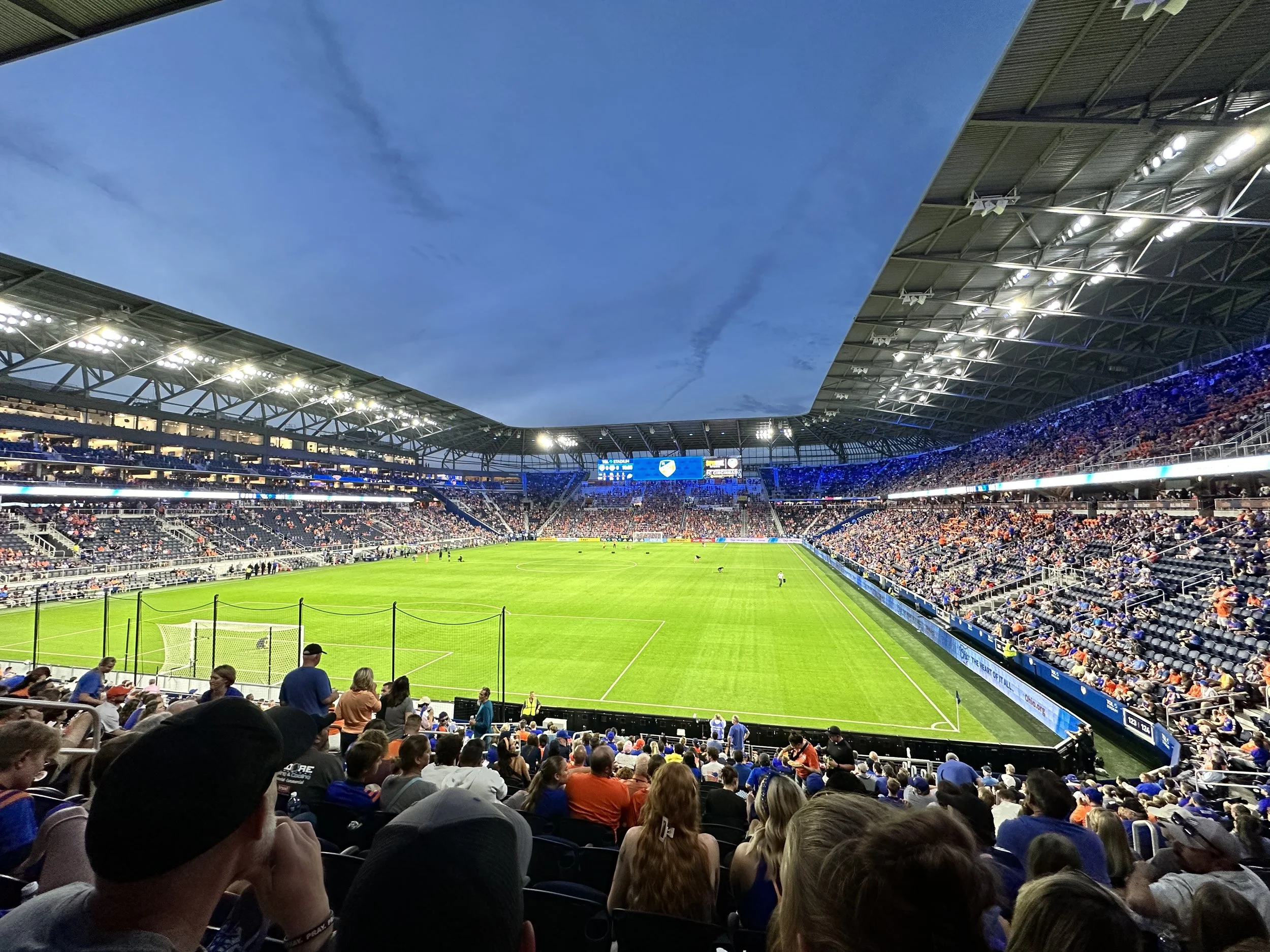 A large stadium filled with spectators watching a soccer game under a partially open roof during the evening, with the sky visible through the opening.
