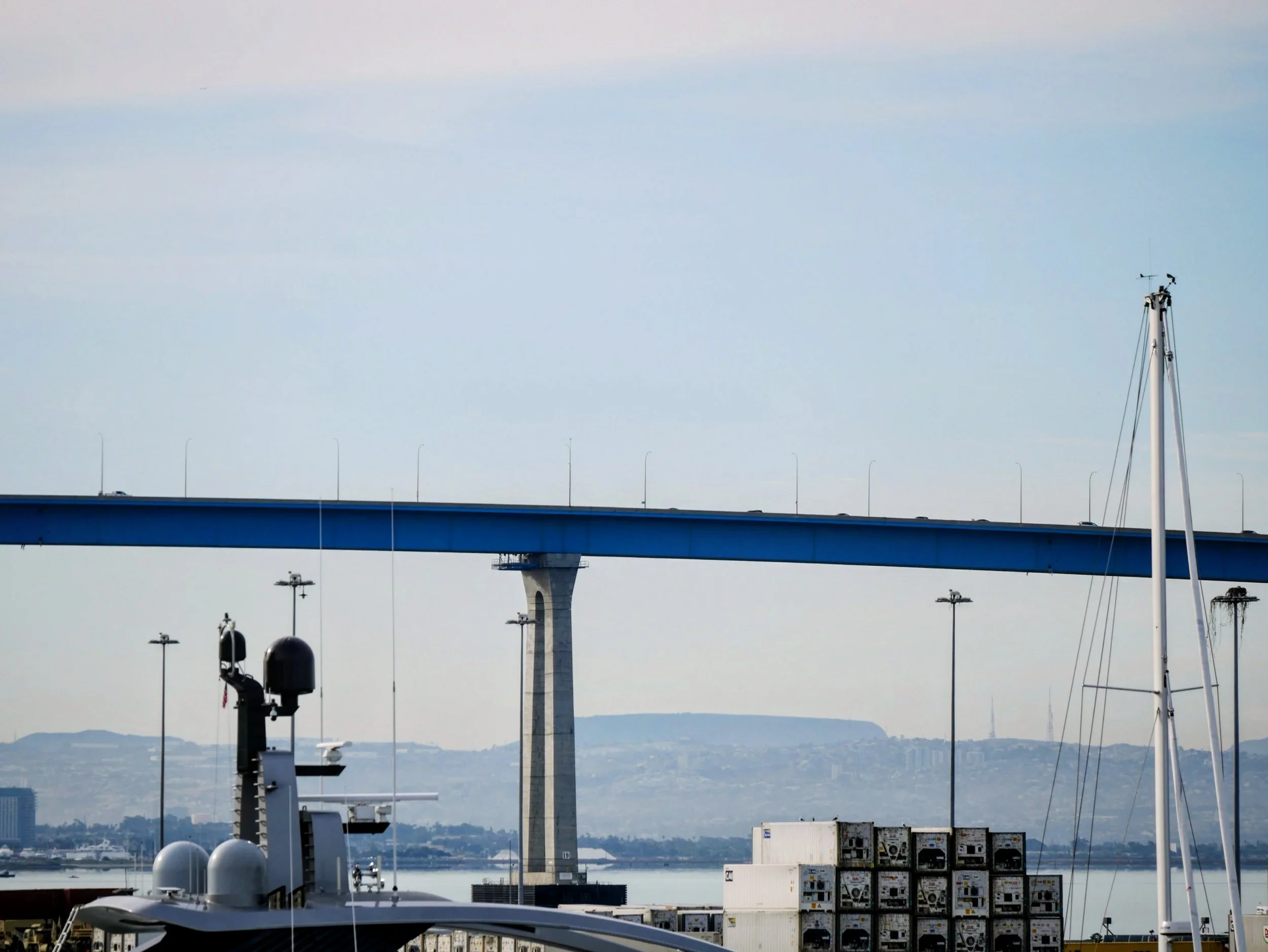 View of a blue bridge over water with a boat and shipping containers in the foreground, and hills in the background under a cloudy sky.