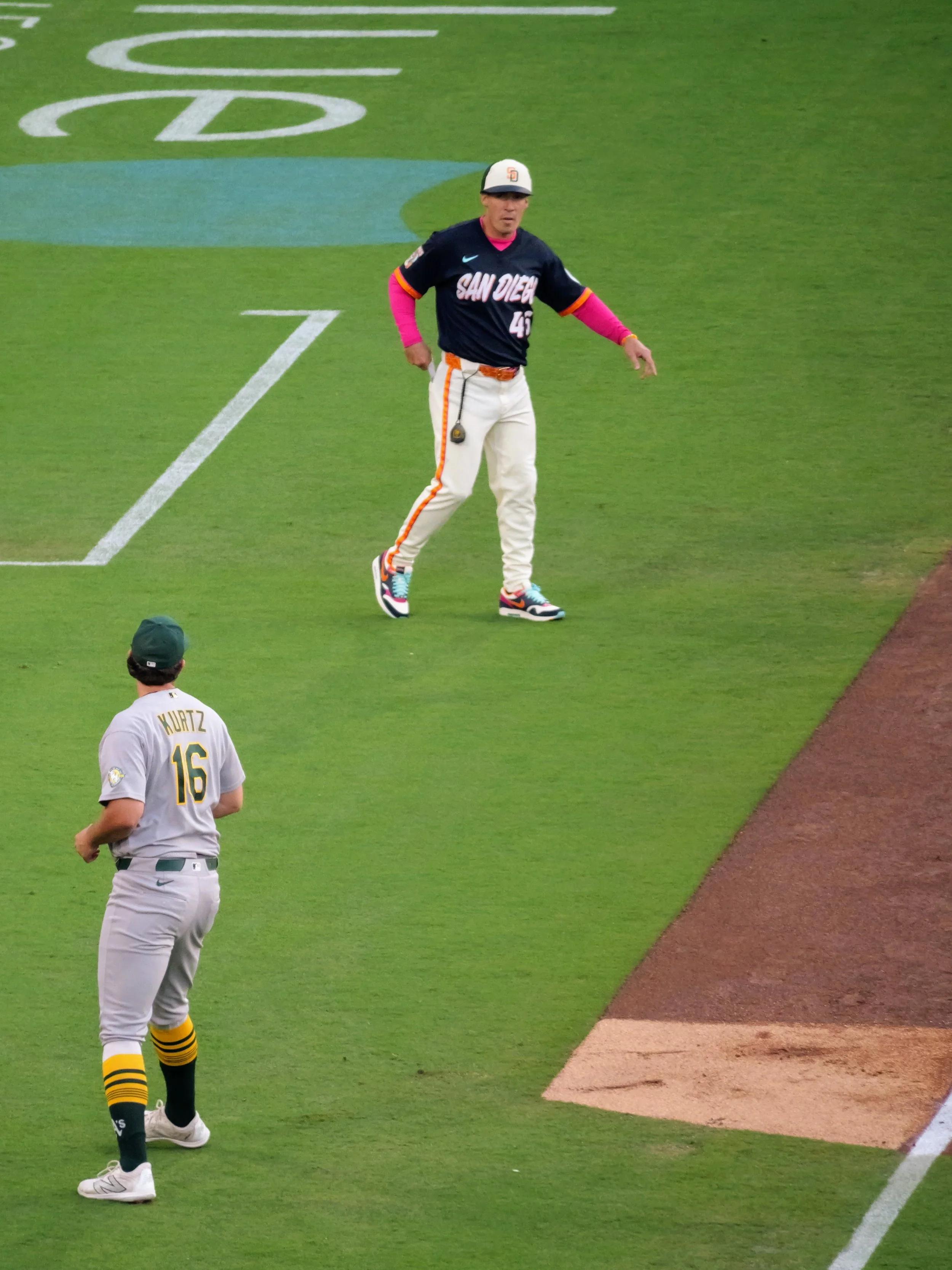 A baseball player in a black jersey with 'SAN DIEGO' written on it, white pants with orange and black stripe, pink sleeves, and colorful sneakers standing on the field. Another player in a gray uniform with yellow and black striped socks and a green 