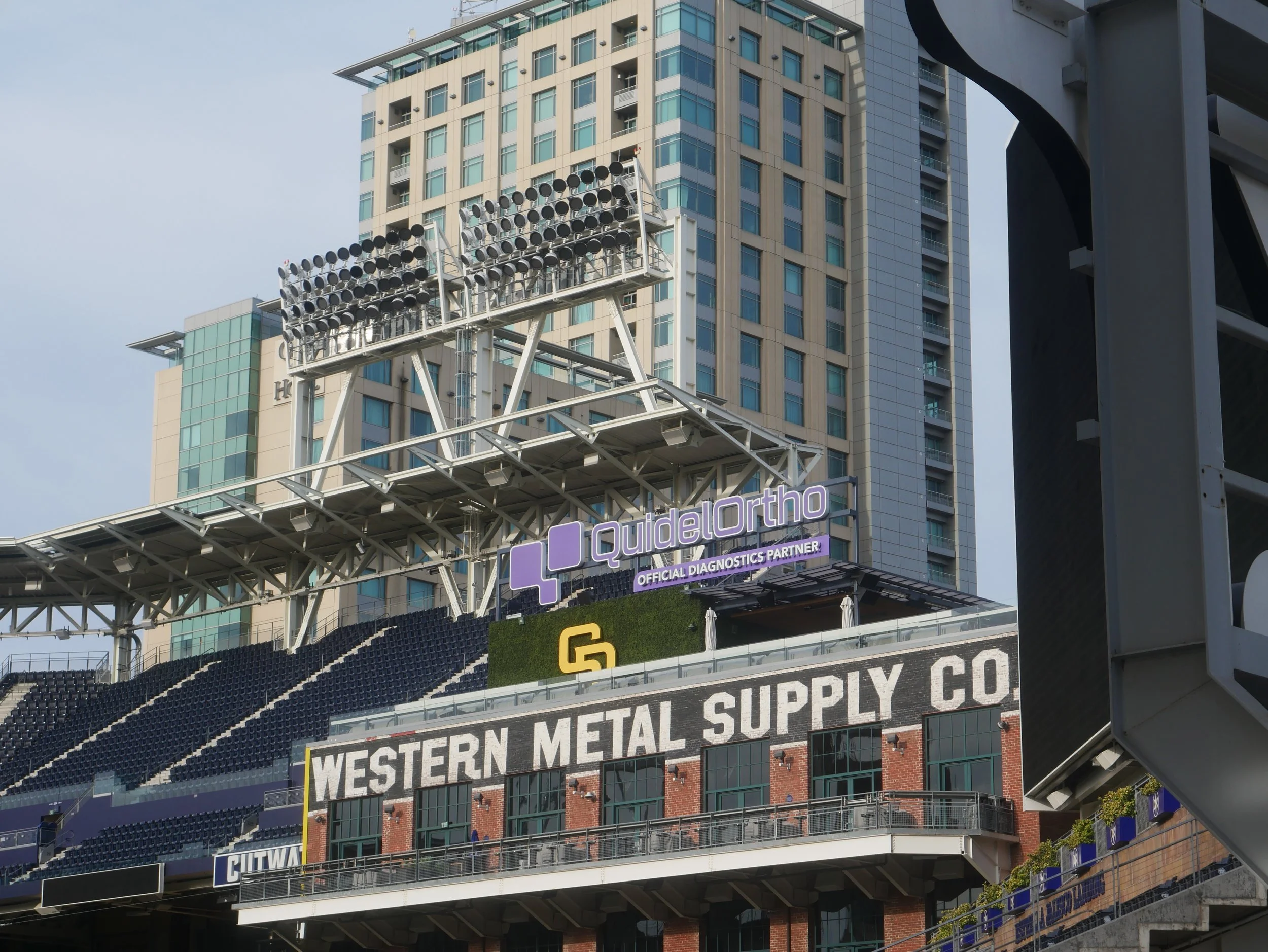 Part of a stadium with a large sign reading 'WESTERN METAL SUPPLY CO' and another digital board with 'QdelOrtho' advertising, with an urban high-rise building behind.