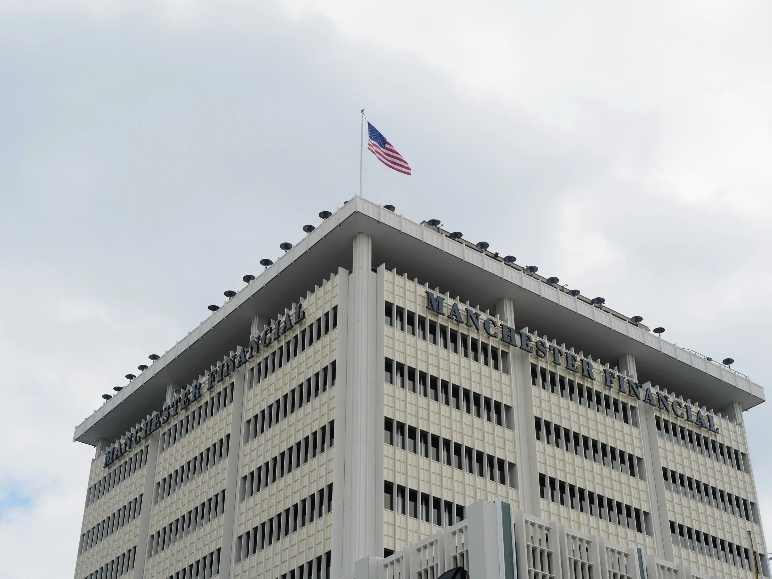 A tall modern office building with a white facade and multiple rows of windows, with the name partially visible. An American flag is flying from the building's rooftop against a cloudy sky.