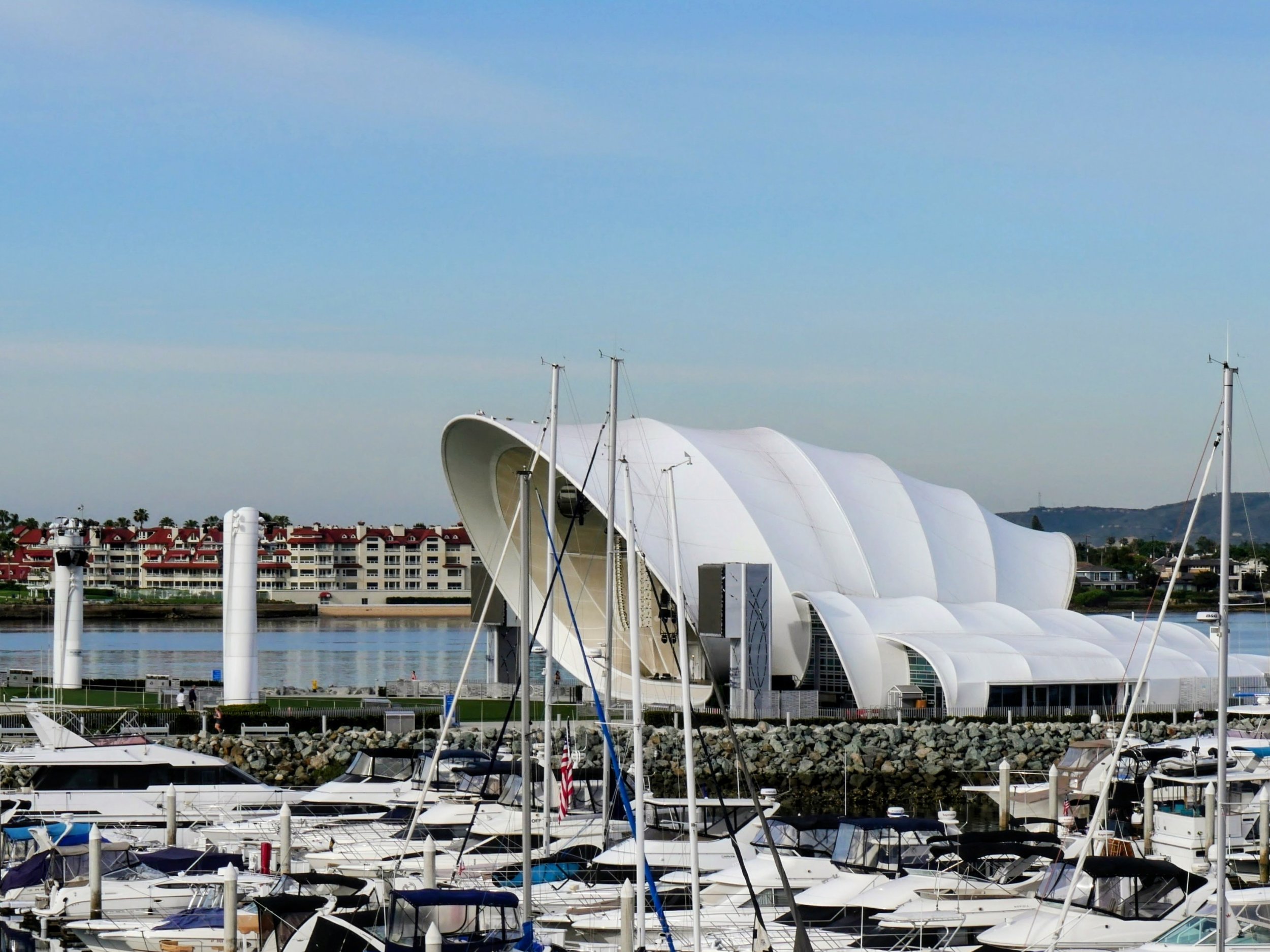 Marina with docked boats in the foreground and a modern, unique white building with curved roof structures on the water's edge.
