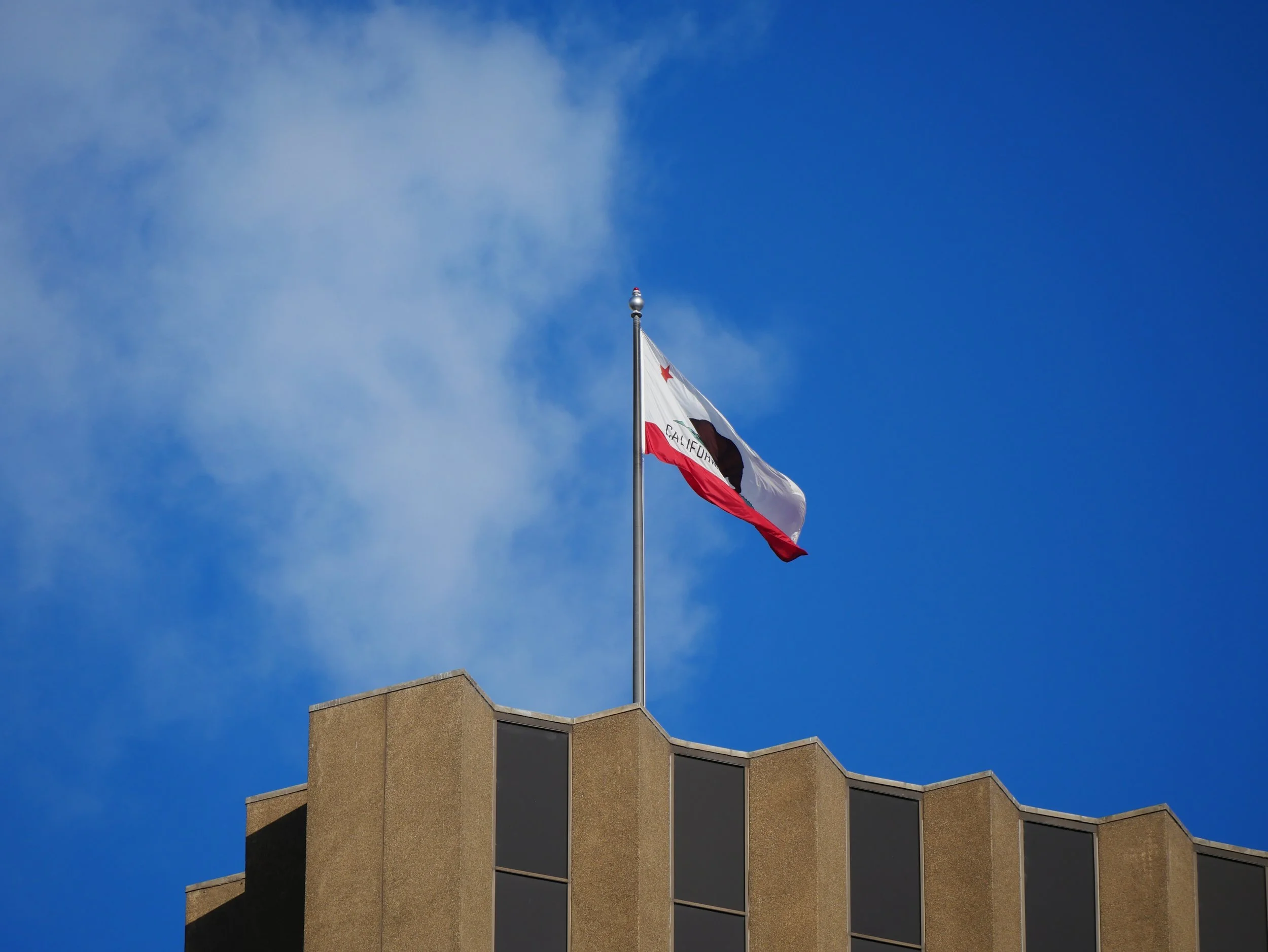 The California state flag flying atop a building against a blue sky with some clouds.