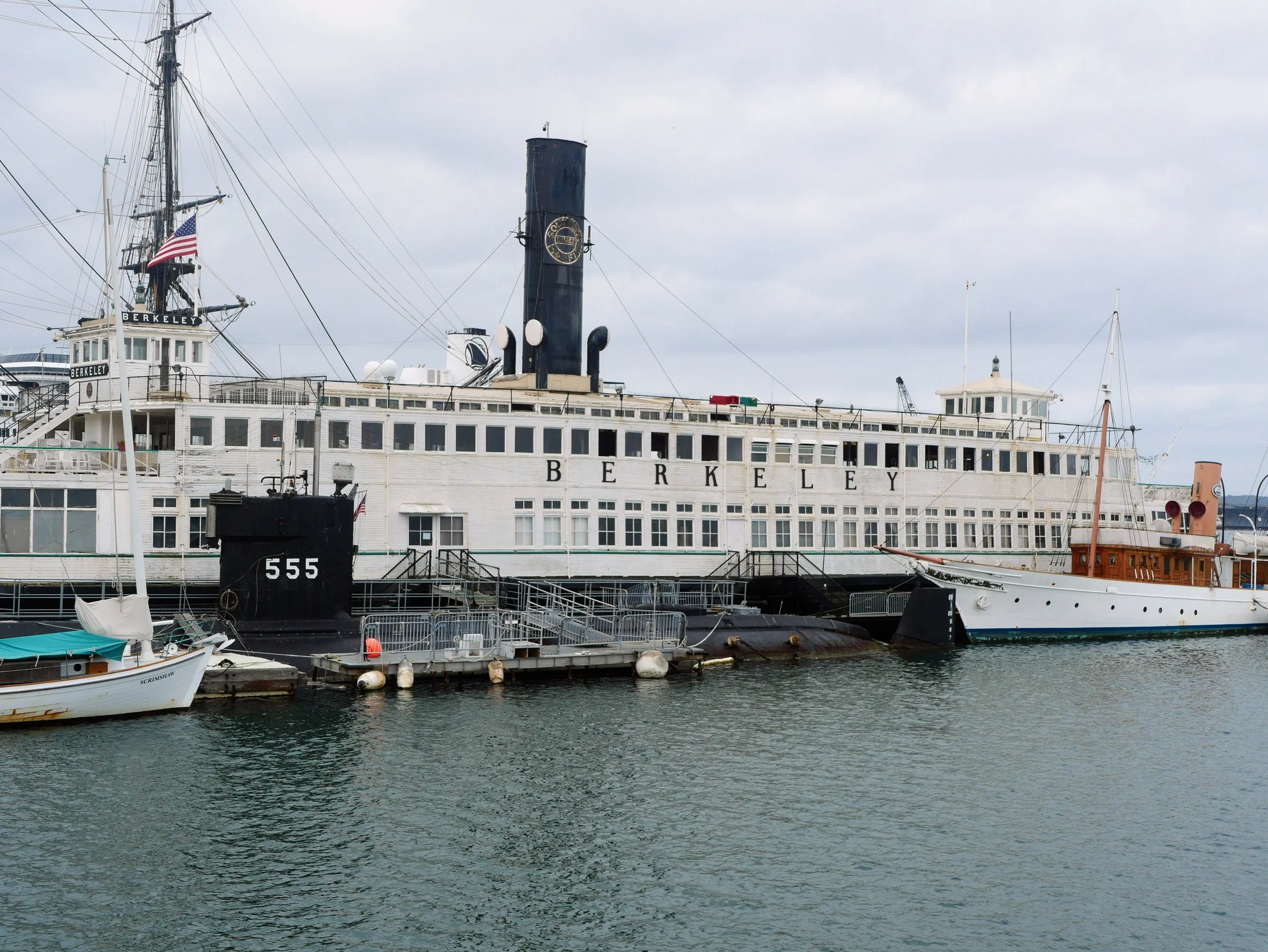 A large white cruise ship with the name 'Berkeley' docked at a marina, with a black smokestack, masts, and multiple decks. Smaller boats are moored in front of it, and the water is calm under a cloudy sky.