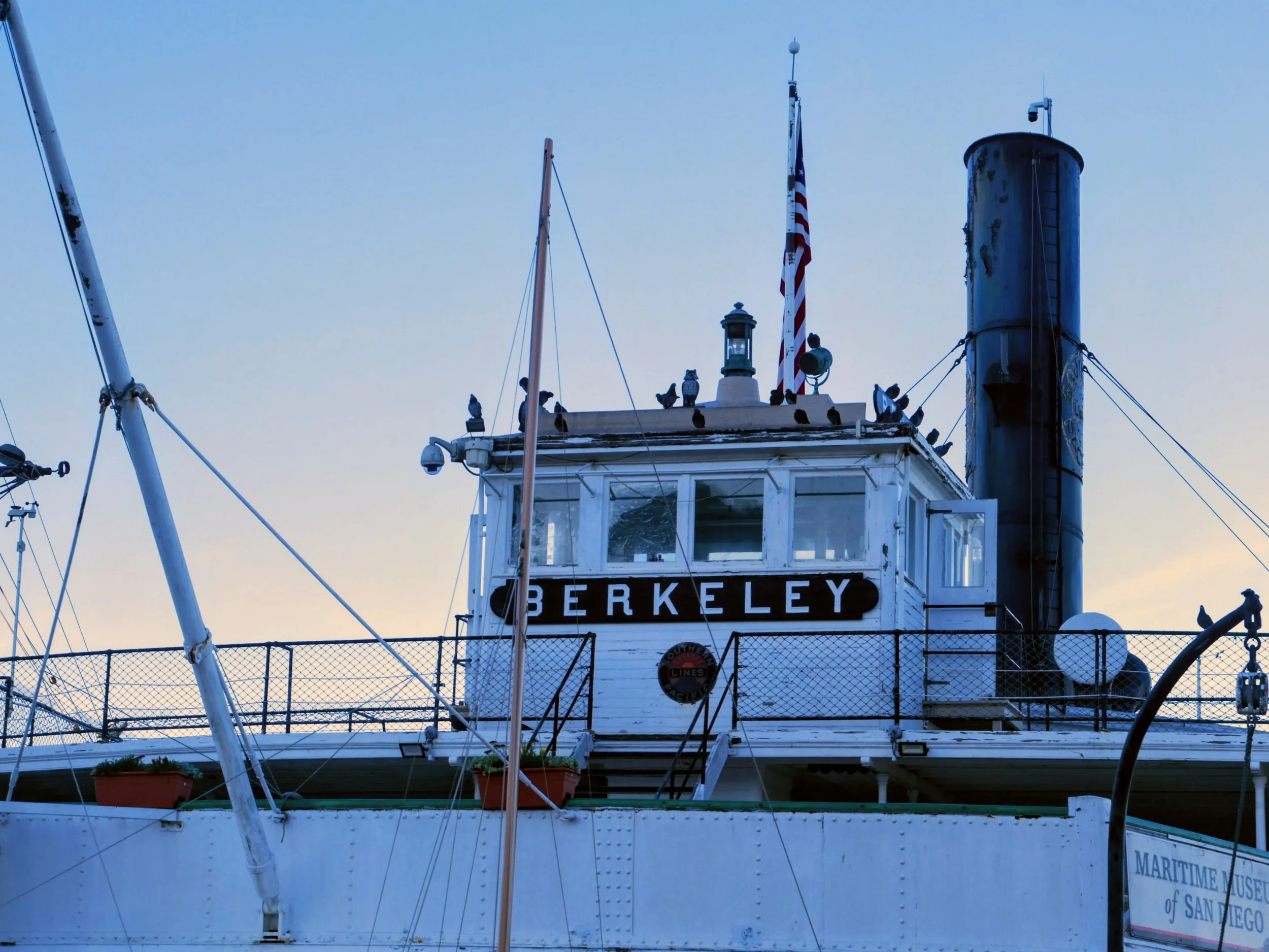 A vintage boat named Berkeley docked at a marina, with pigeons on the roof, American flag, and a clear sky in the background.