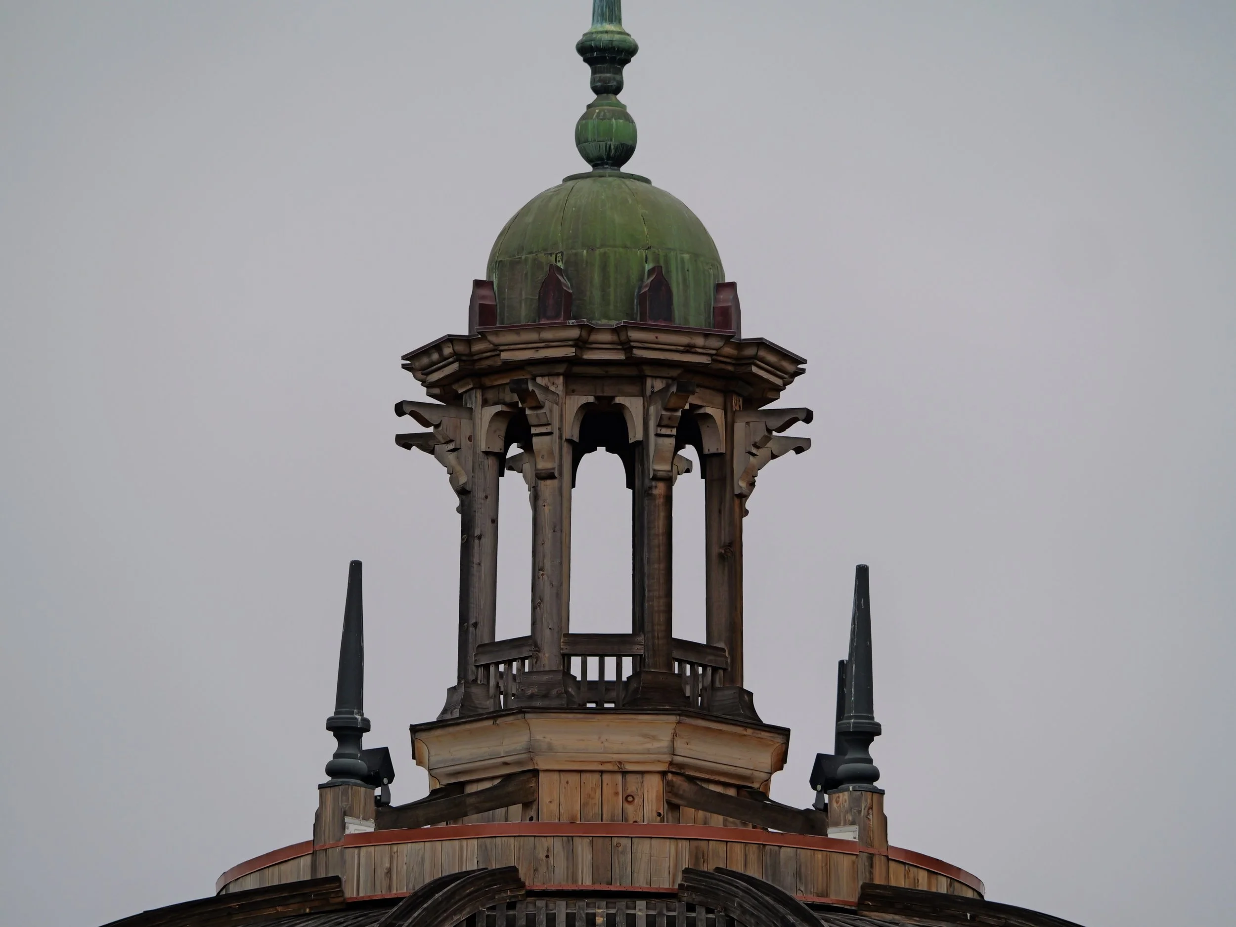 The top of a wooden tower with a green dome and decorative elements, flanked by two black spire-like structures, against a gray sky.