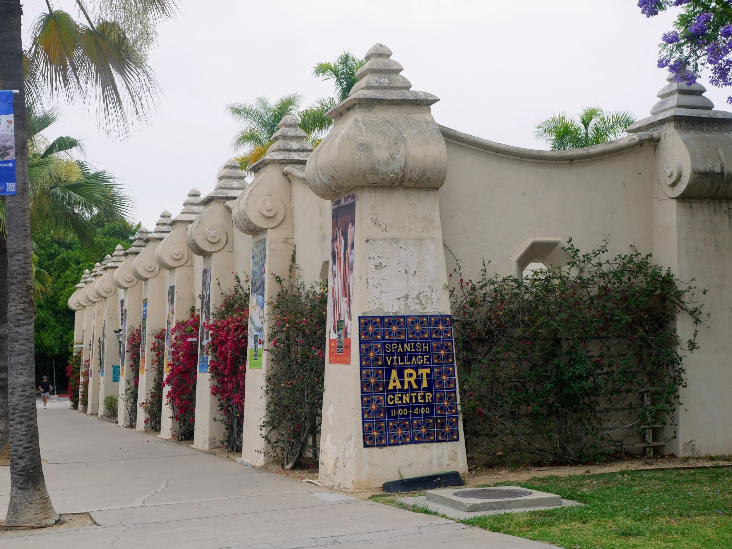 A beige wall with decorative columns topped with scrolls and pointed finials, adorned with posters and a sign for an art center, surrounded by palm trees and flowering bushes on a cloudy day.