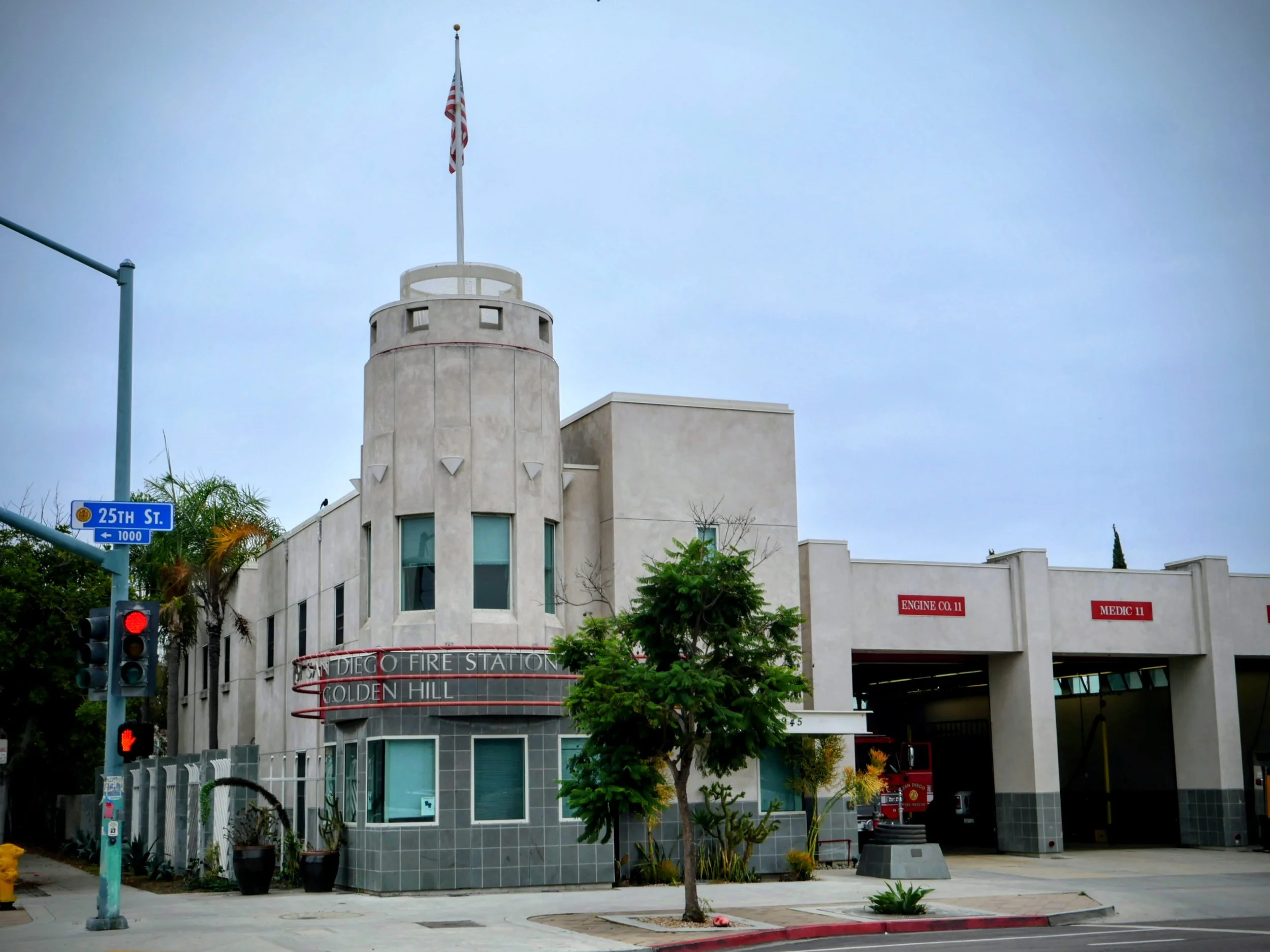 Facade of the San Diego Fire Station with a circular tower, American flag on top, street sign reading '25th St.', and trees in front.