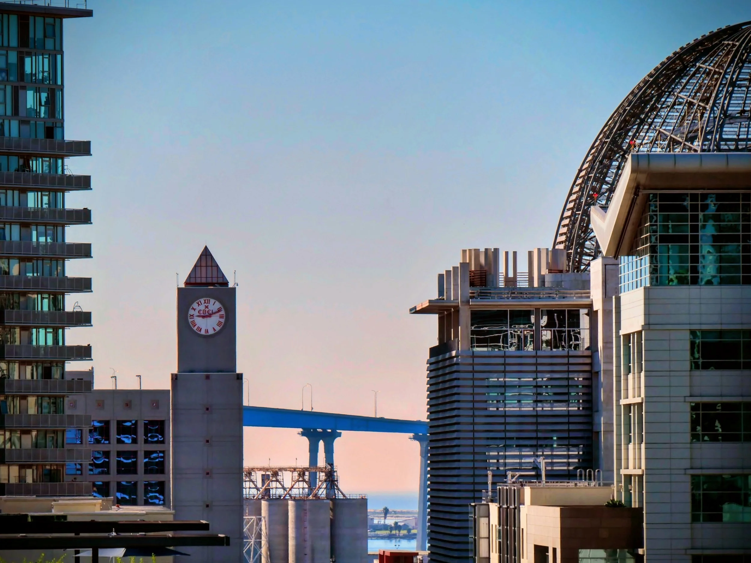Cityscape of modern buildings with a clock tower in the foreground and a bridge in the background, under a clear sky.