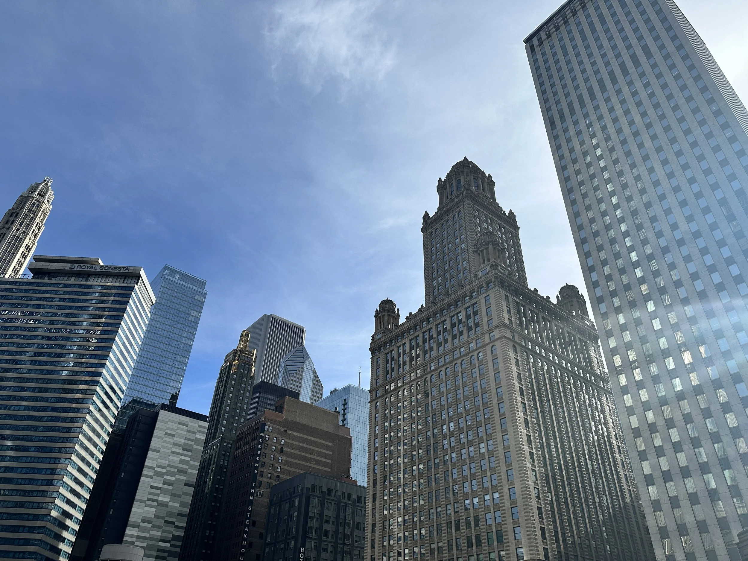 A cityscape view looking up at tall skyscrapers against a blue sky with some clouds. The buildings include modern glass structures and historic stone and brick buildings.