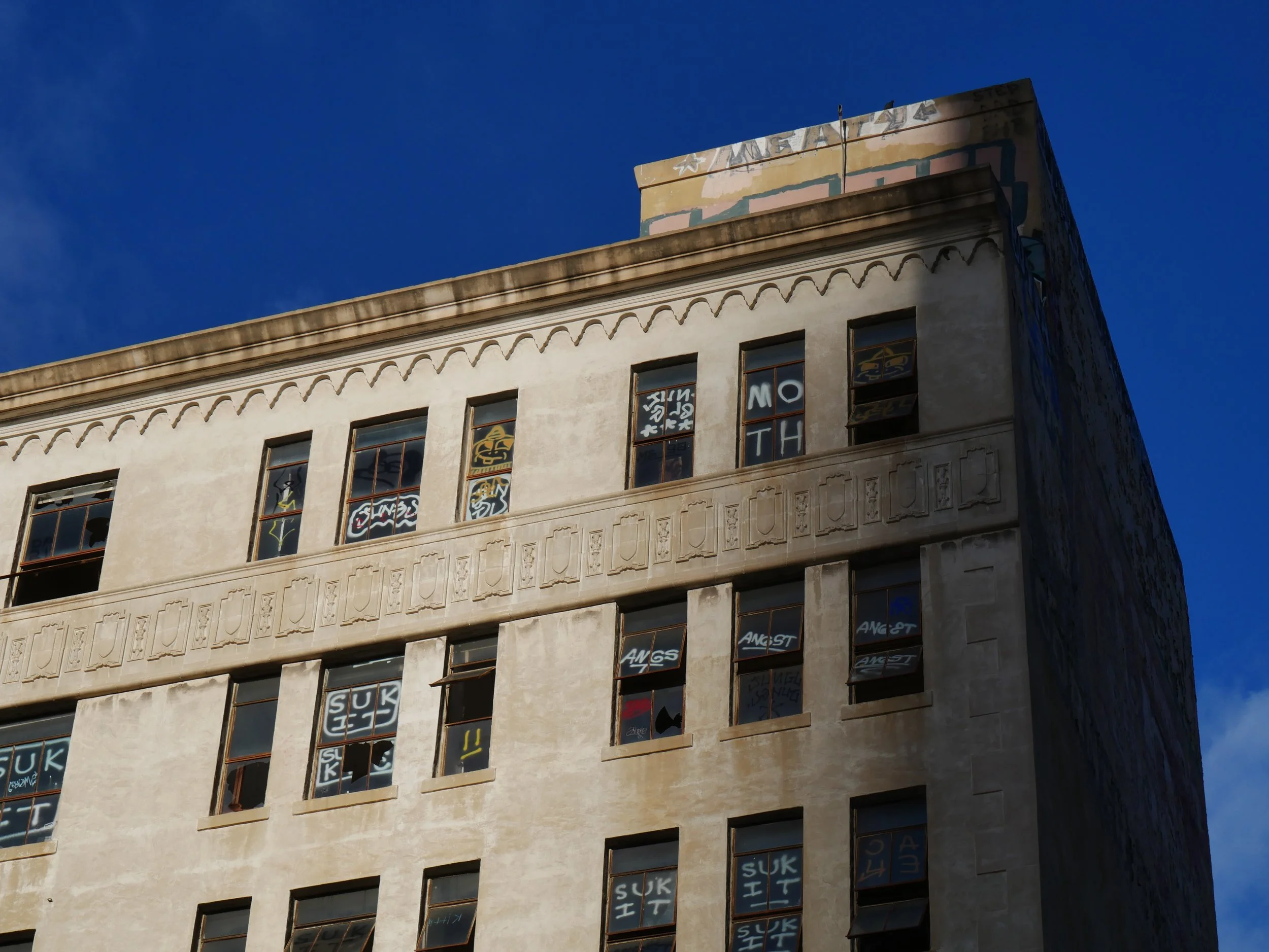 The top of a beige building with several tall narrow windows displaying various messages written on papers in the windows, against a clear blue sky.