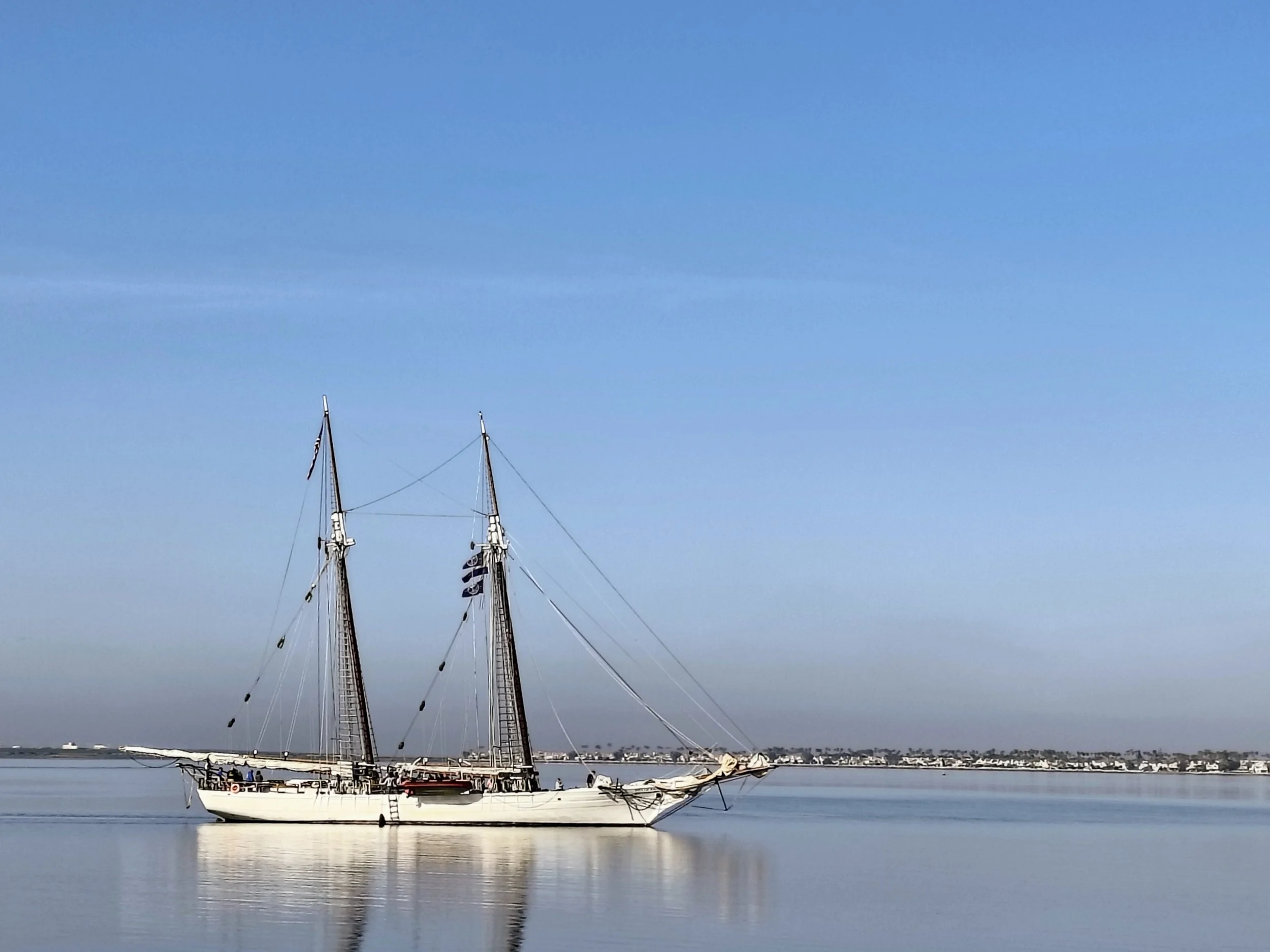 Sailboat with two masts floating on calm water near the shore, with a distant cityscape under a clear blue sky.