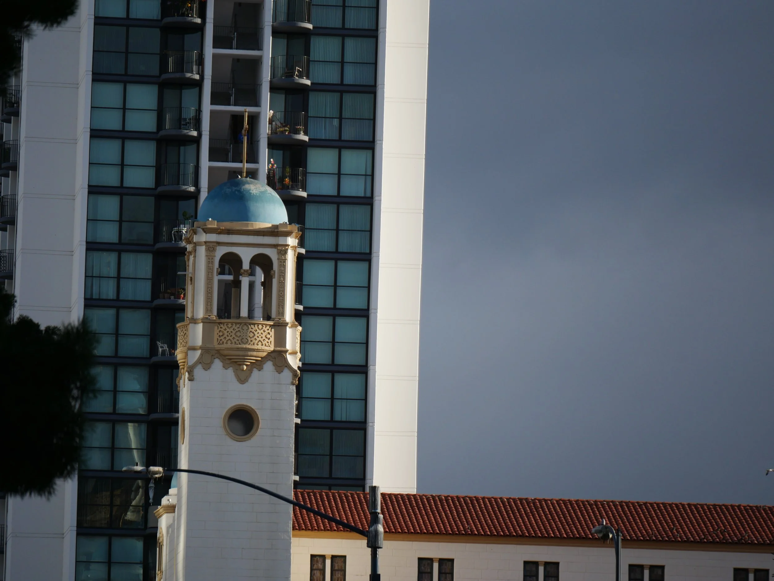 A tall modern building with glass windows and small balconies, next to a white tower with a blue dome and rooftop decorations. Power lines and a streetlamp are in the foreground, with an overcast sky in the background.