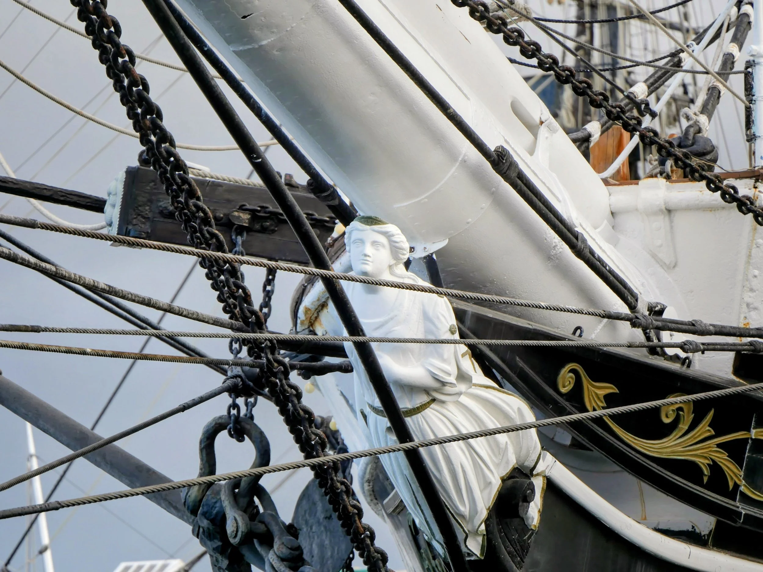 Close-up of the bow of a sailing ship featuring a white decorative figurehead of a woman, surrounded by black and bronze chains, with rigging and ropes around it.