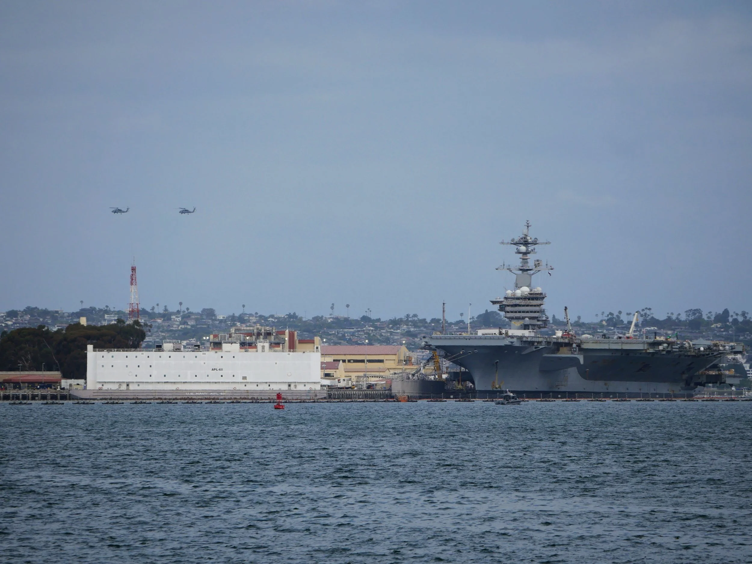 A view of the water with a large naval ship docked near the shoreline. In the sky, two helicopters fly in formation near a city skyline.